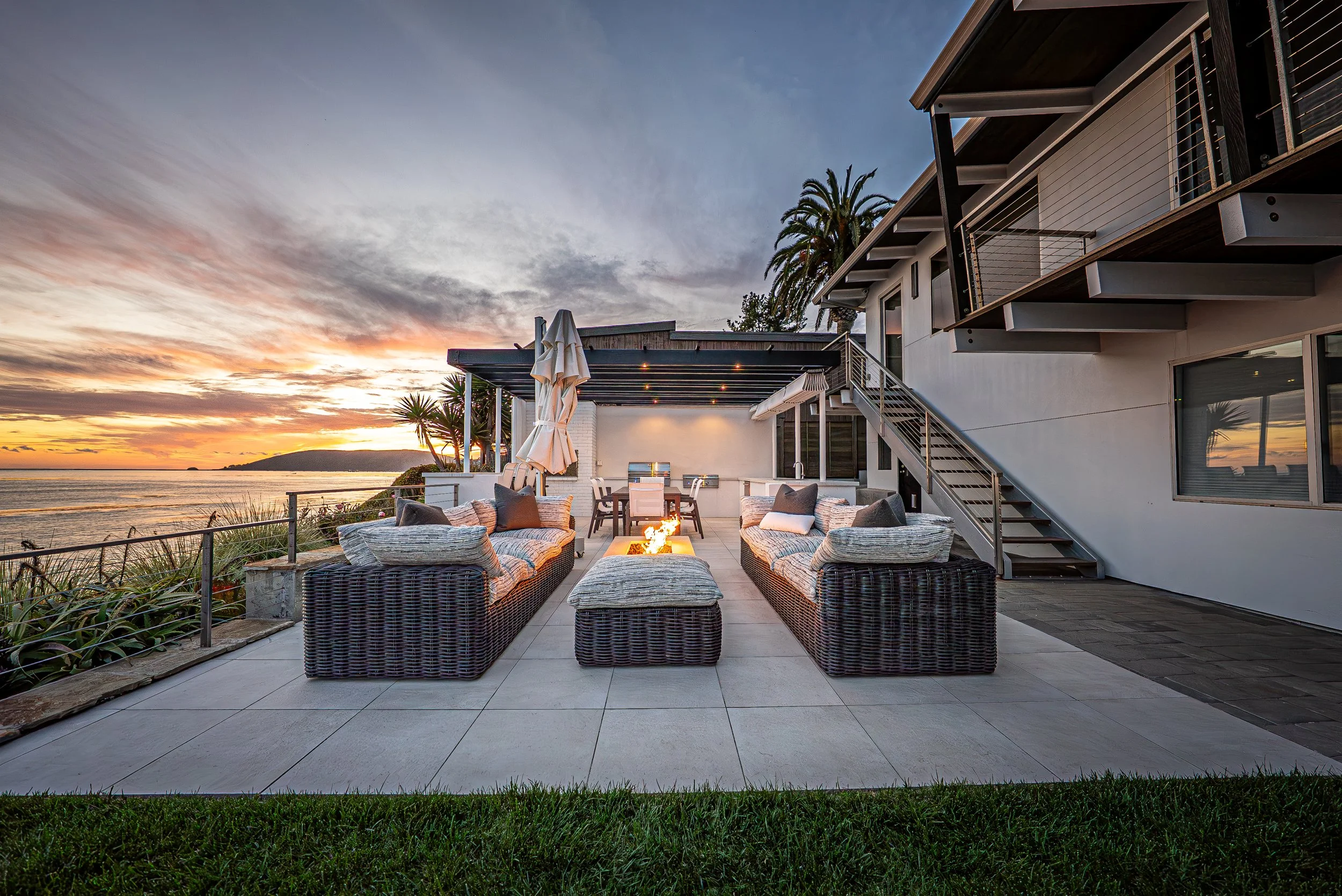 Outdoor patio with wicker furniture, fire pit, dining area, and view of the ocean at sunset