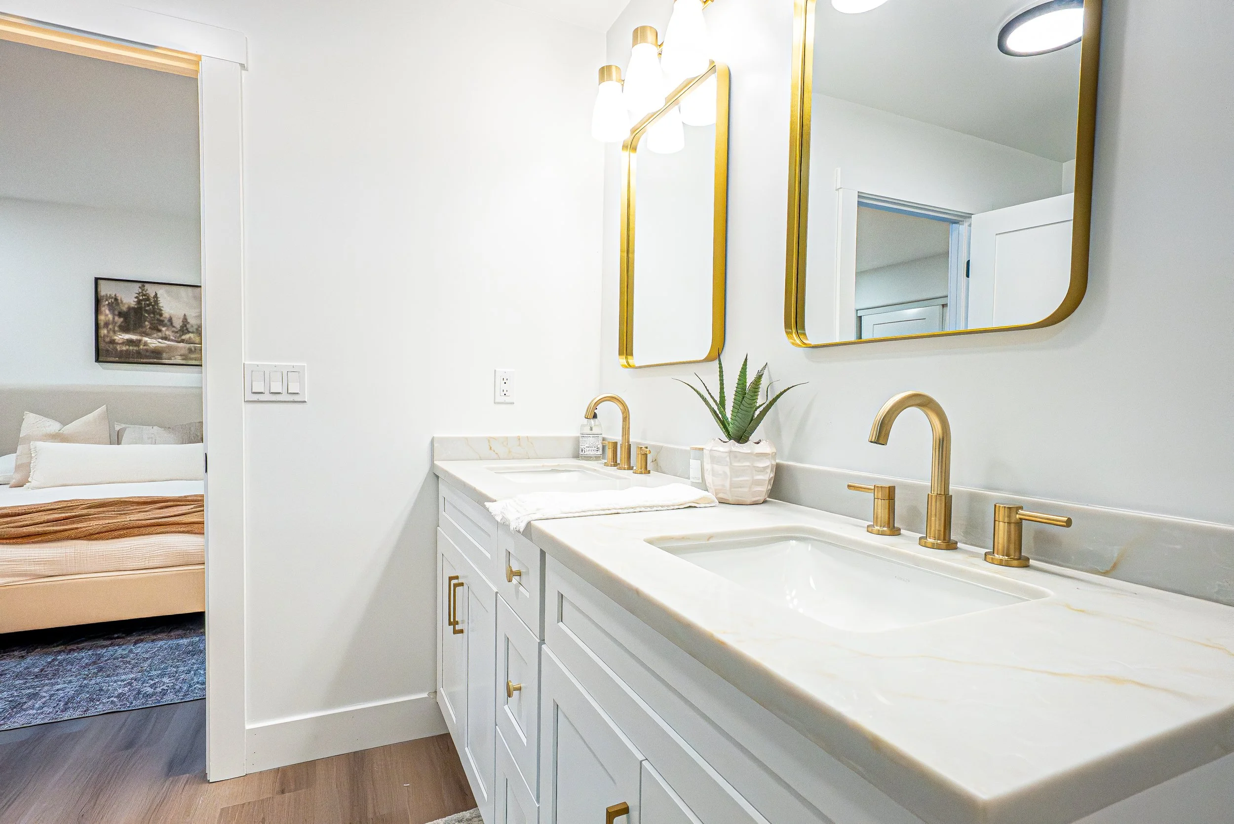 Modern bathroom with white vanity, gold fixtures, two mirrors, and a potted plant. Bedroom visible in the background.