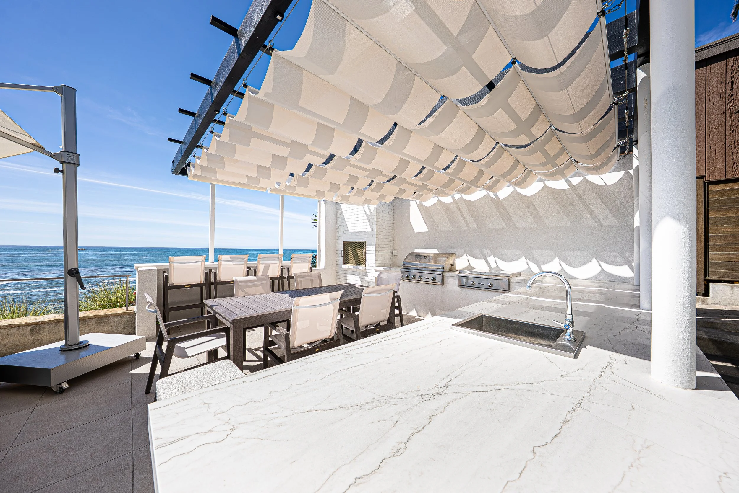 Outdoor terrace with barbecue grill, dining table with chairs, and ocean view, shaded by a beige canopy.