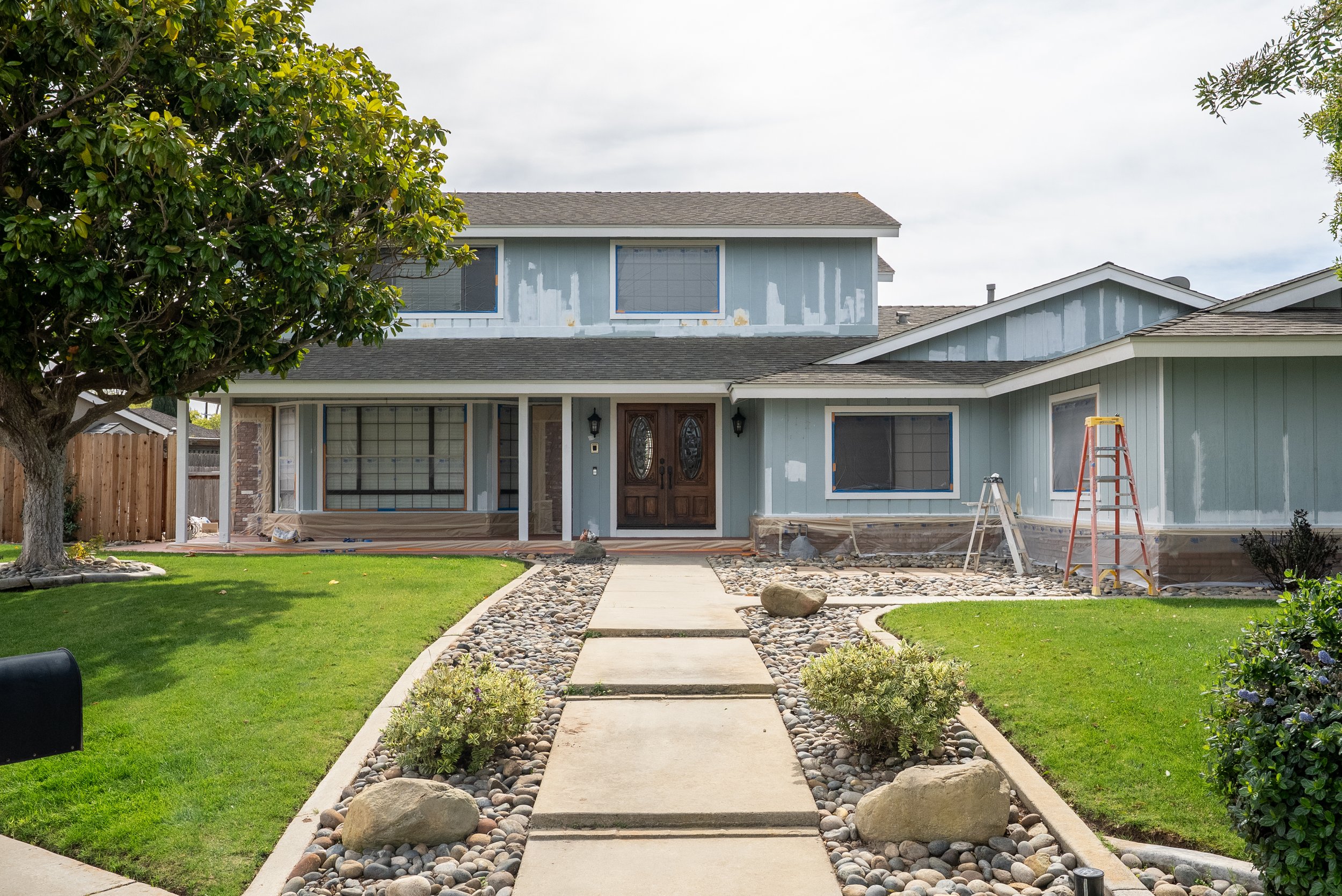 A two-story house under renovation, with painted blue exterior walls, a front yard with a concrete walkway, green grass, trees, and rocks, and construction ladders and tools near the house.