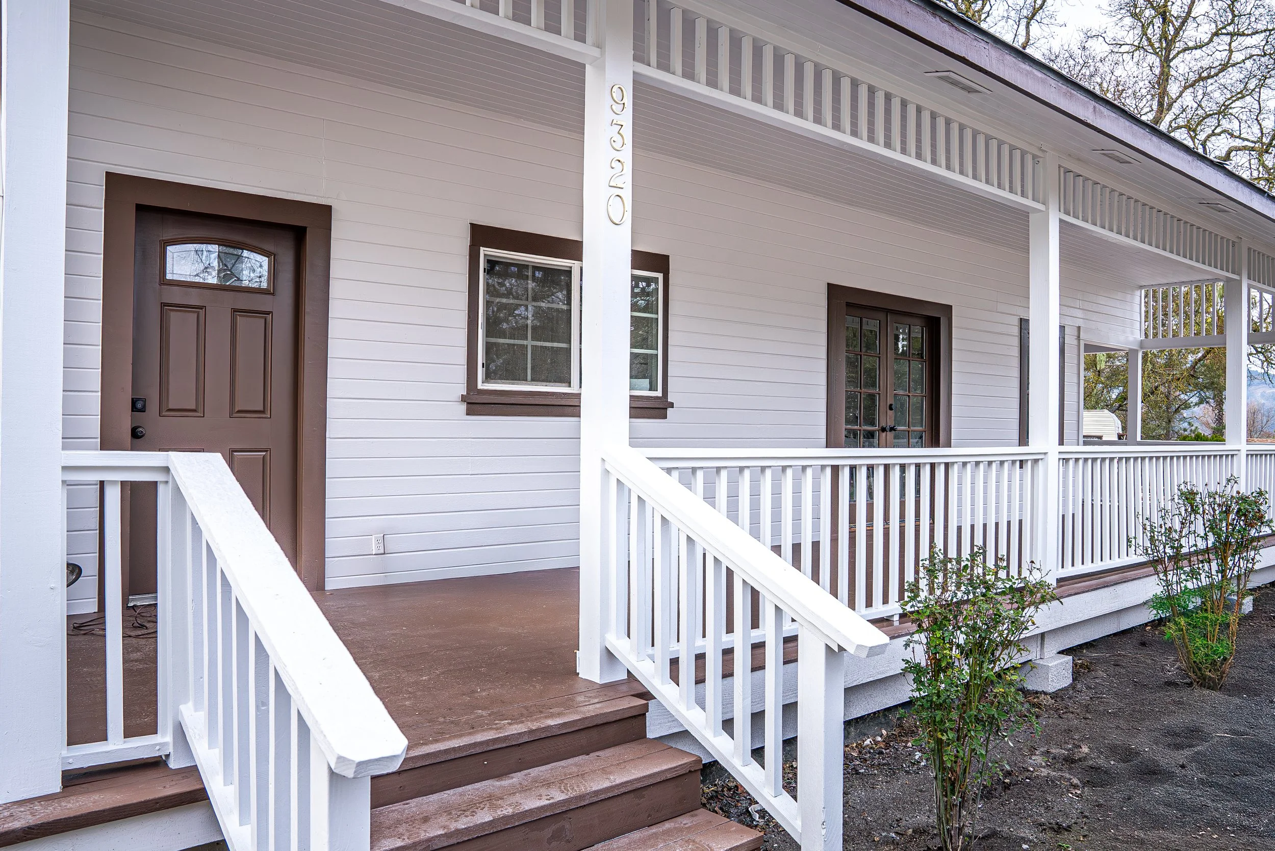 Front porch of a house with white siding, brown wooden steps, a brown front door, a window, and a glass door. The house number 9320 is visible on a white post.