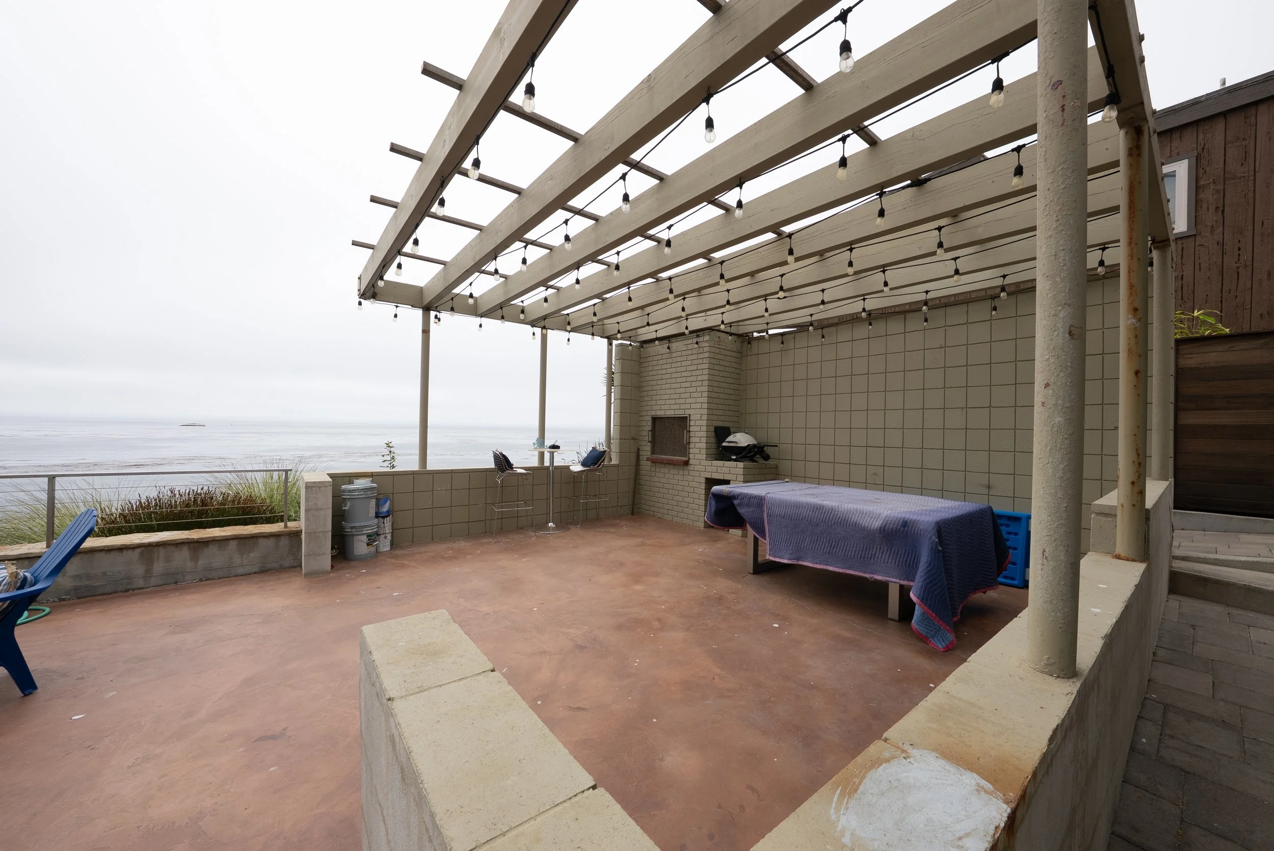 Outdoor patio with string lights, a table covered with a blue tablecloth, a small grill, two chairs, and a view of the ocean in the background.
