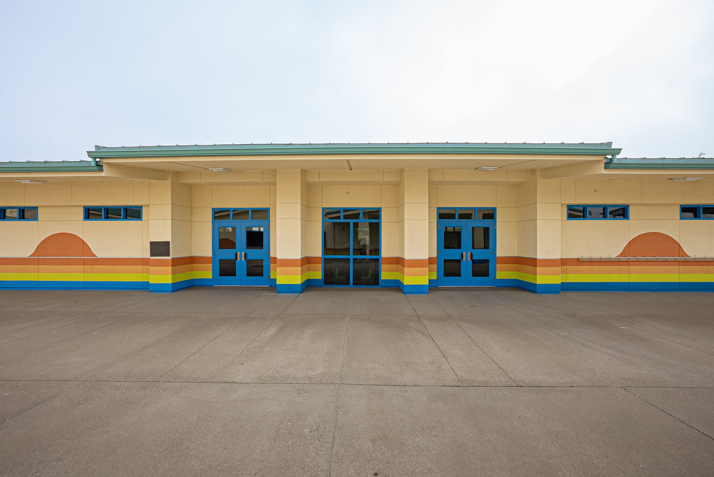Facade of a colorful building with blue doors and rainbow stripe along the wall, and a paved sidewalk in front.