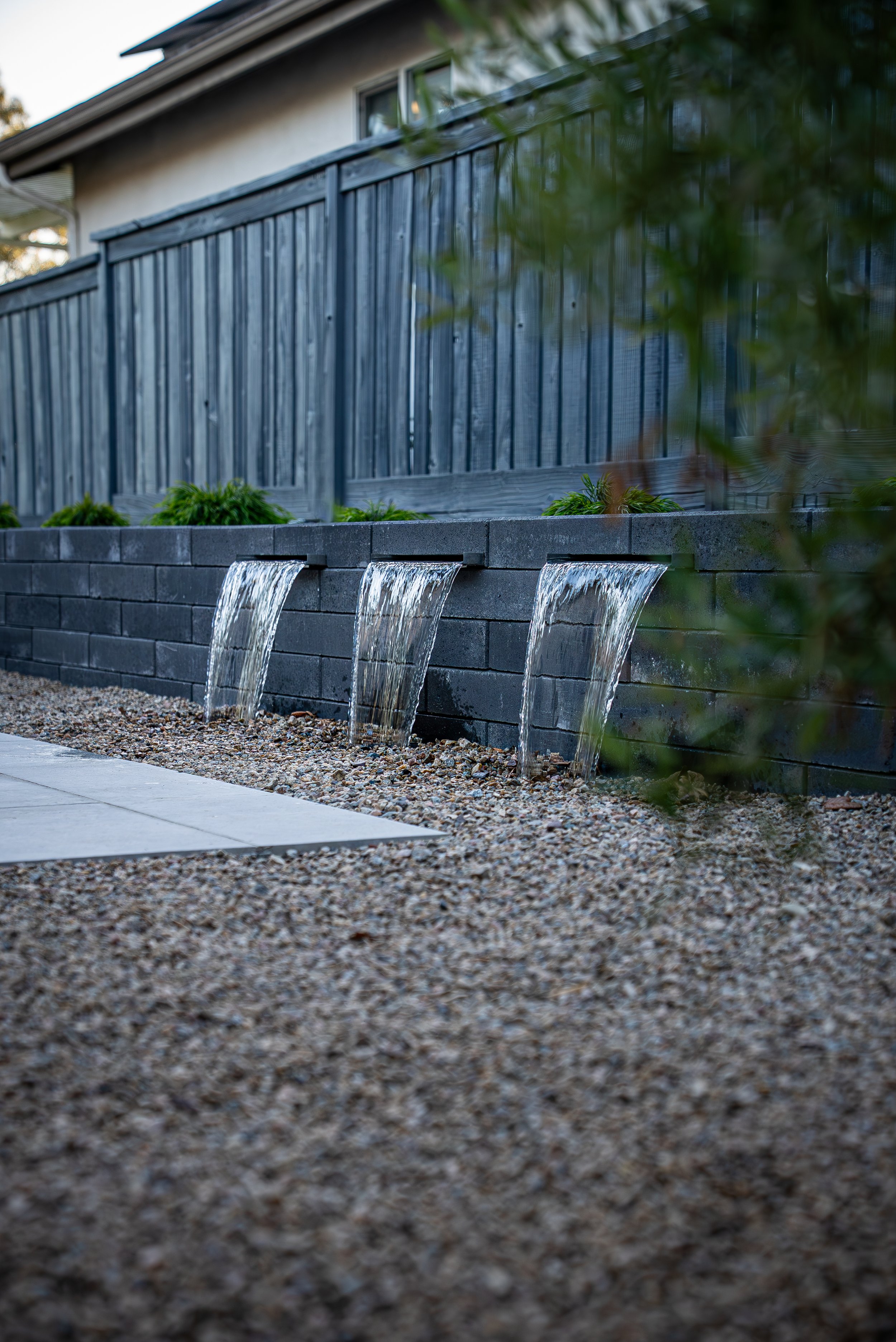 Exterior backyard scene with a black stone retaining wall and three small waterfalls flowing into a gravel area, with a grey wooden fence and a house in the background.