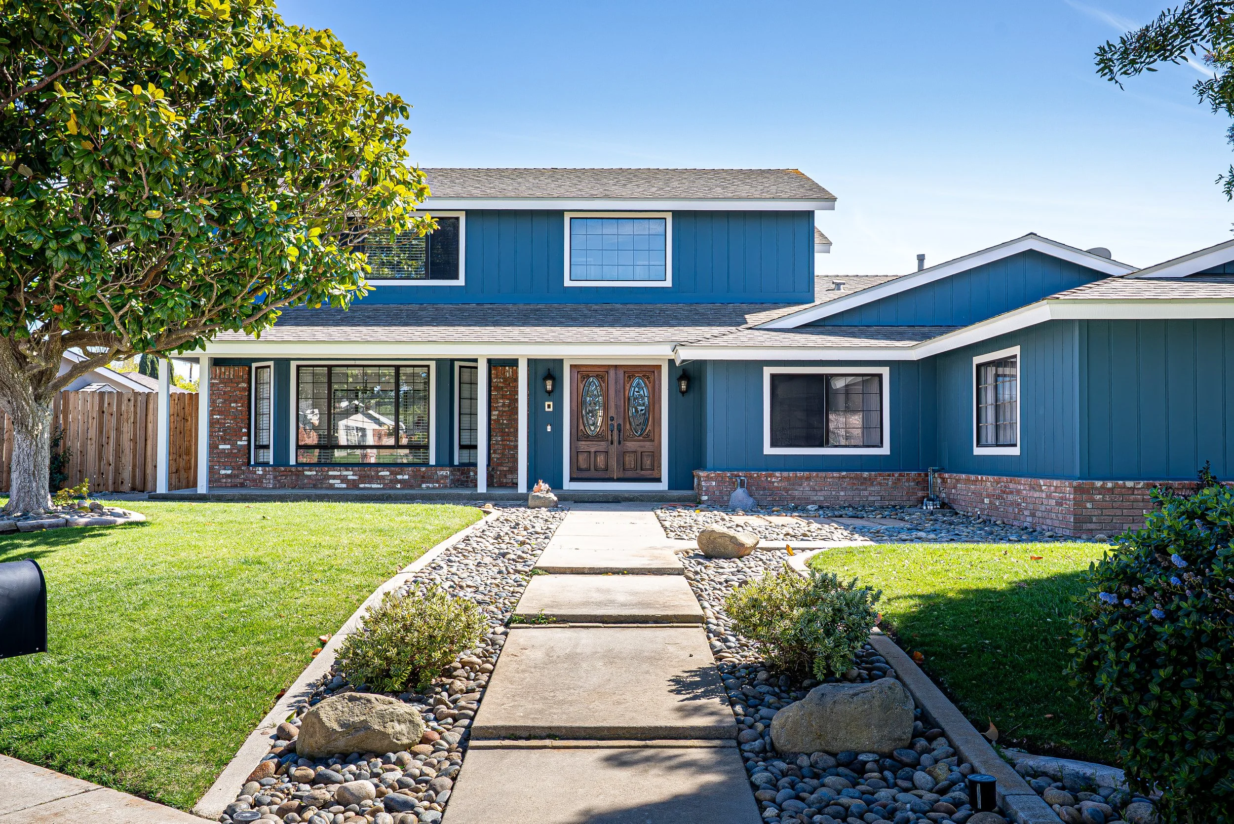 Front view of a two-story house with a blue exterior and brick accents, surrounded by a well-maintained lawn, a stone pathway, and trees under a clear blue sky.