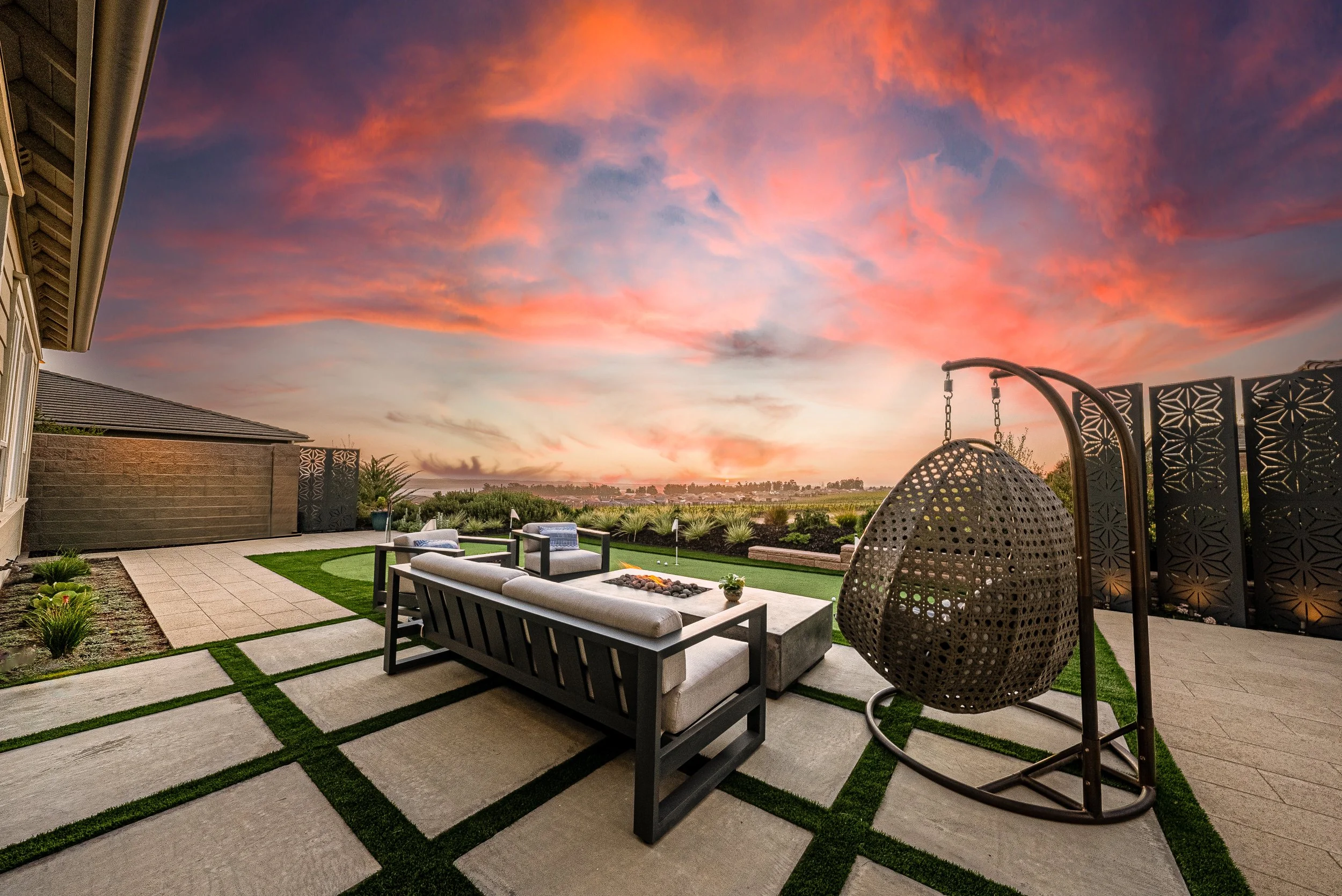 Backyard patio with outdoor furniture, a fire pit, and a hanging egg chair, under a colorful sunset sky.