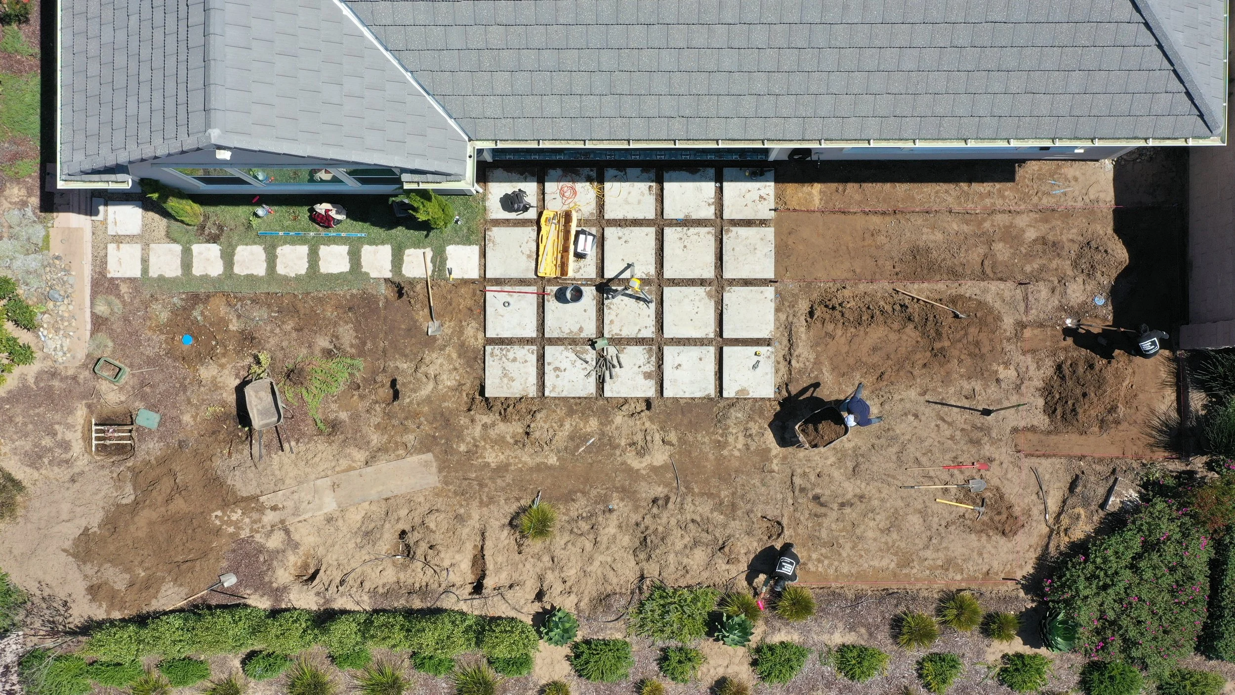 An aerial view of a backyard under construction, with concrete slabs arranged to form a patio area, tools and workers present, dirt and soil areas, and surrounding greenery.