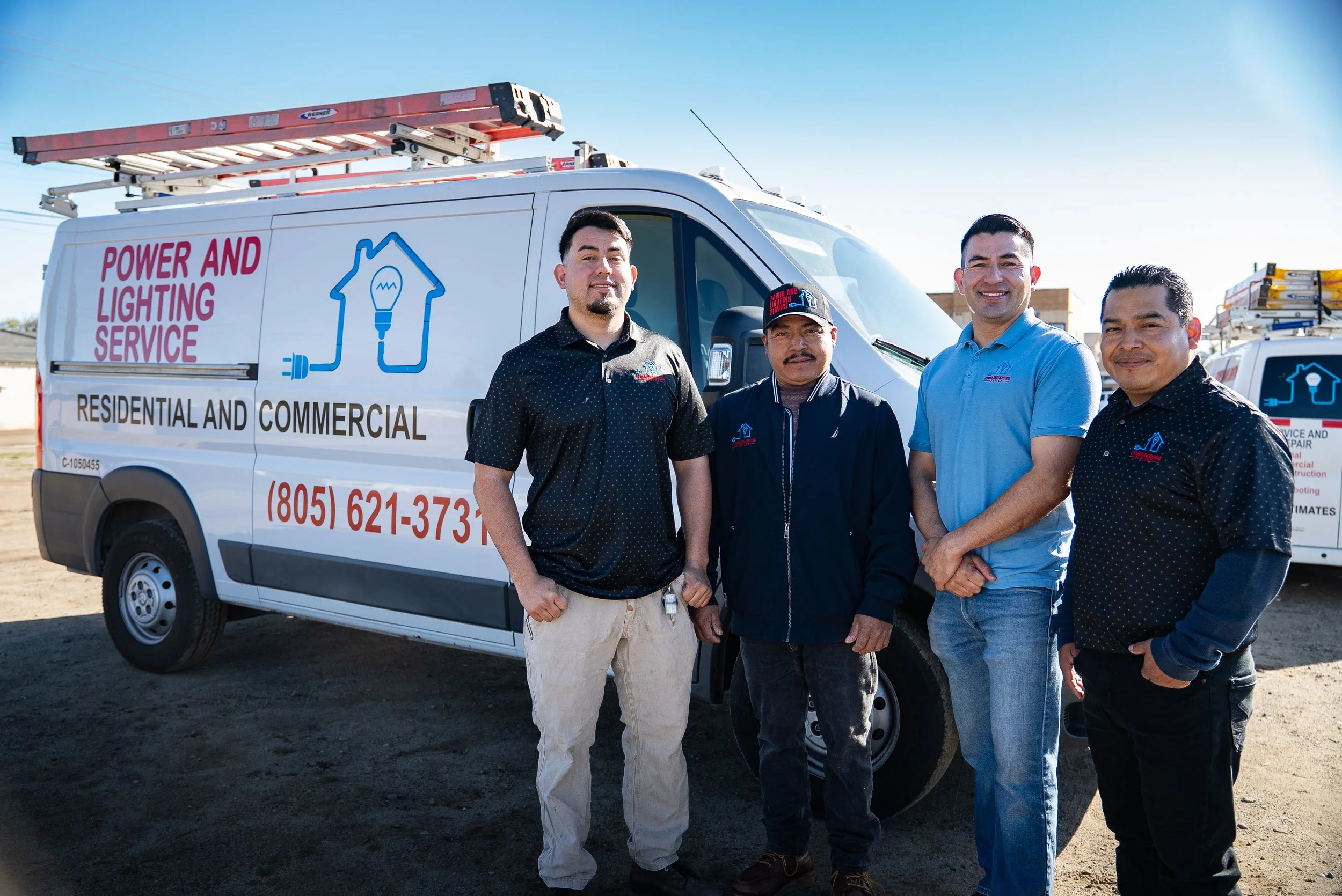 Four men standing in front of a white service van labeled with power and lighting service graphics and contact information. They are outdoors on a clear day.