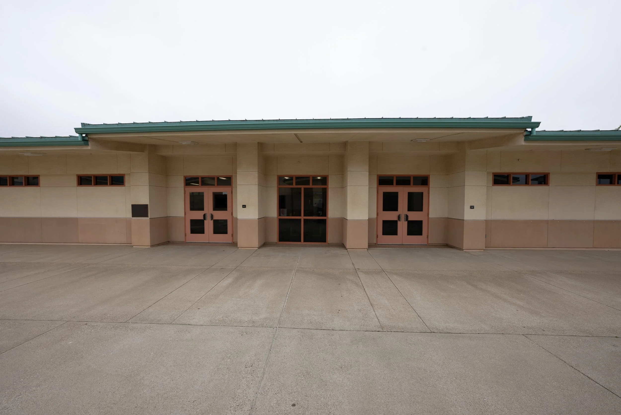 Empty school building with three double doors, large windows, and a green metal roof on a cloudy day