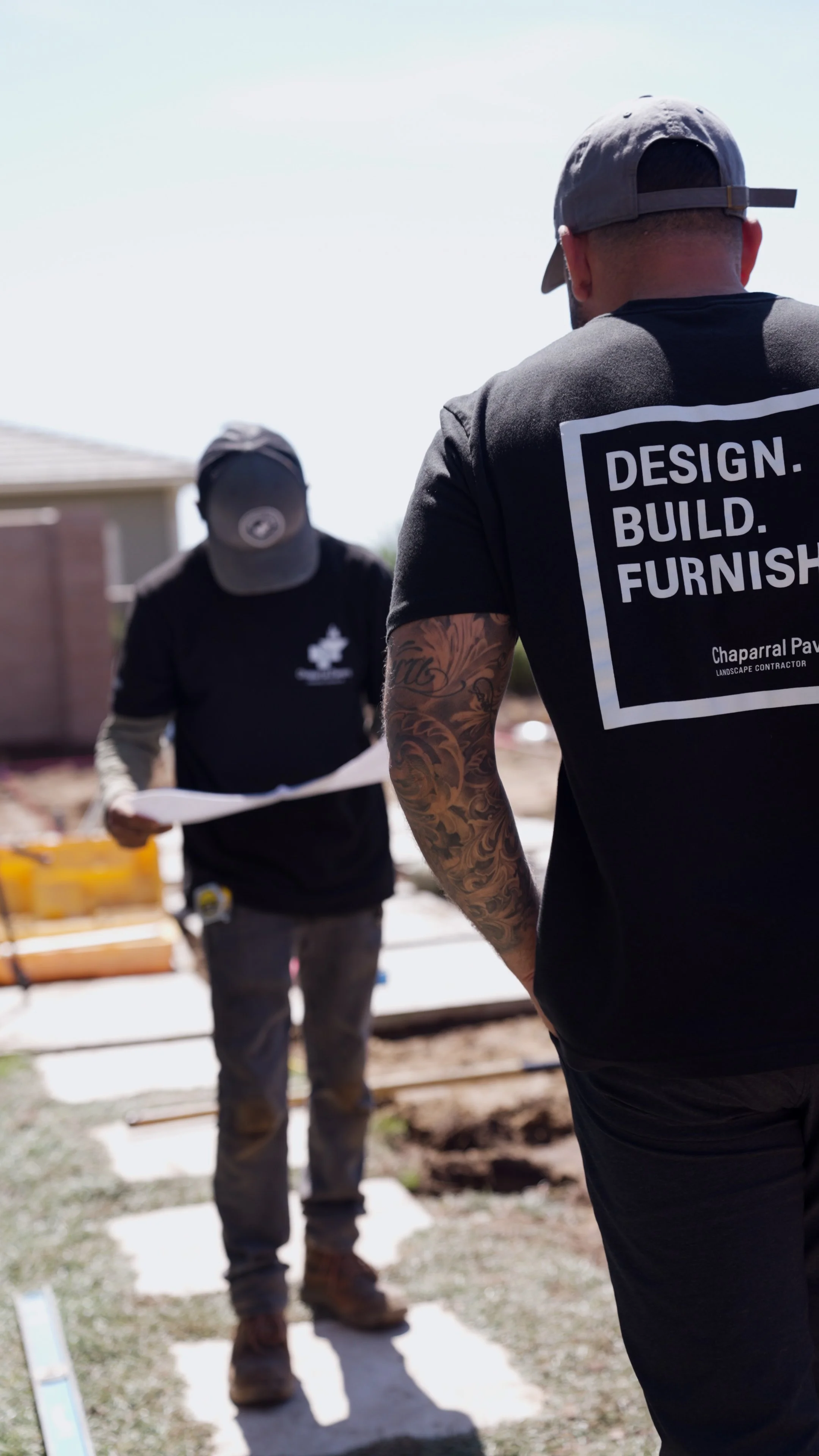 Two construction workers on a building site, one in focus with a tattooed arm, wears a T-shirt saying 'Design. Build. Furnish.' and a baseball cap, the other blurred in the background holding papers, with a house and construction materials behind the