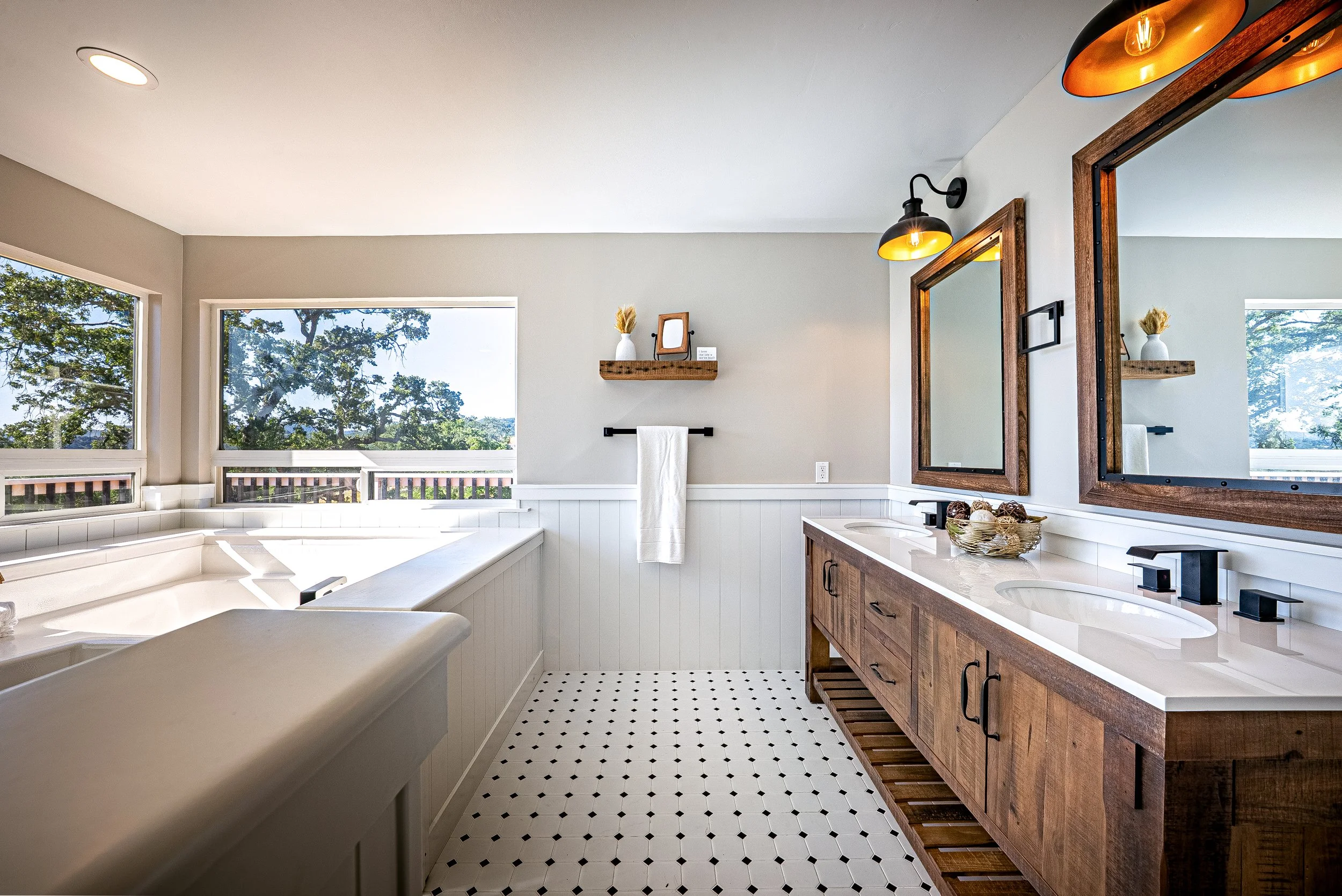 Bathroom with natural light, white beaded wainscoting, a double sink wooden vanity with black hardware, large mirrors, and black light fixtures. A corner bathtub with windows overlooking trees, a shelf with decorative items, and black and white patte