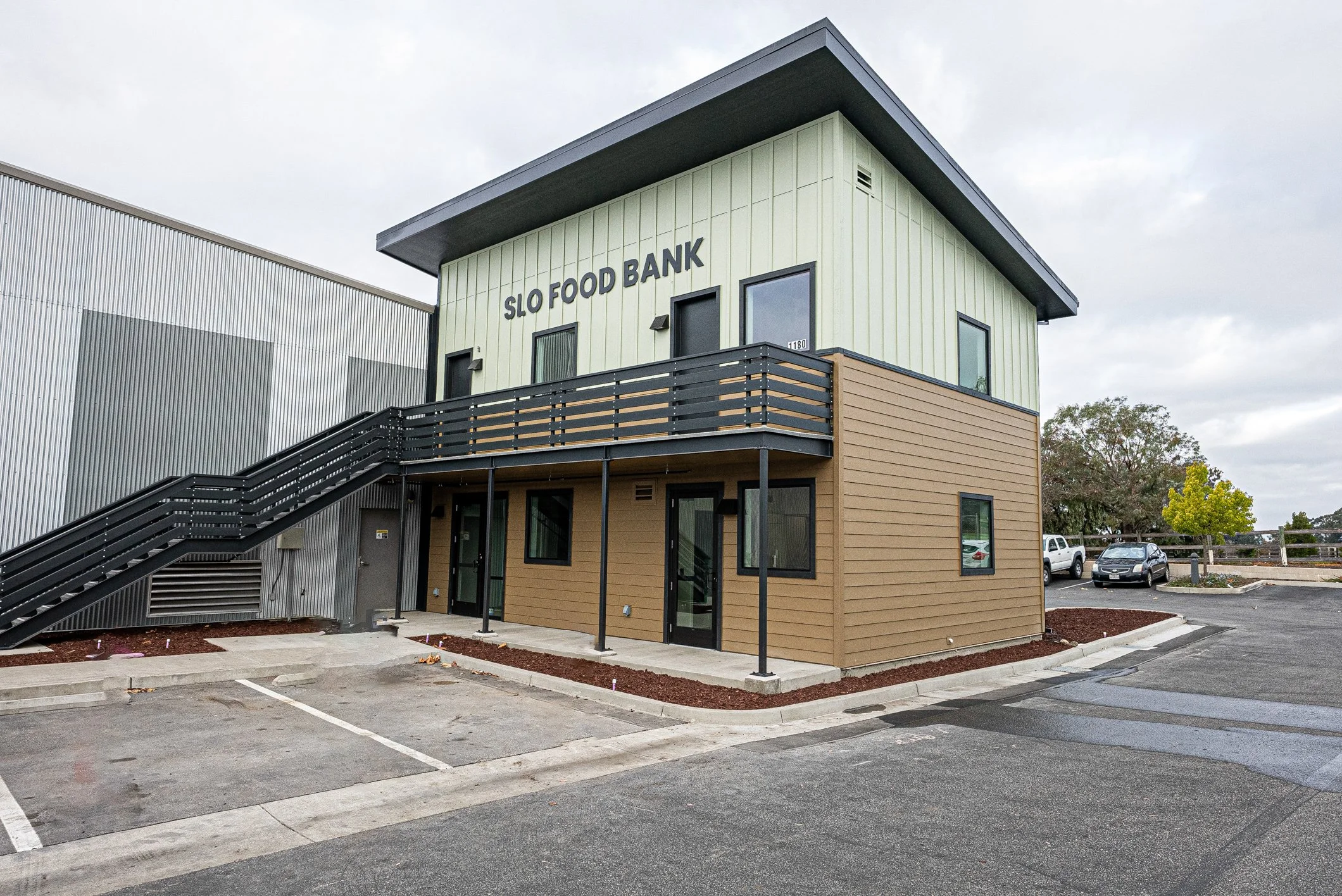 Two-story building labeled "SLO Food Bank" with a black staircase, metal and wood exterior, parking lot in front, and trees in the background.