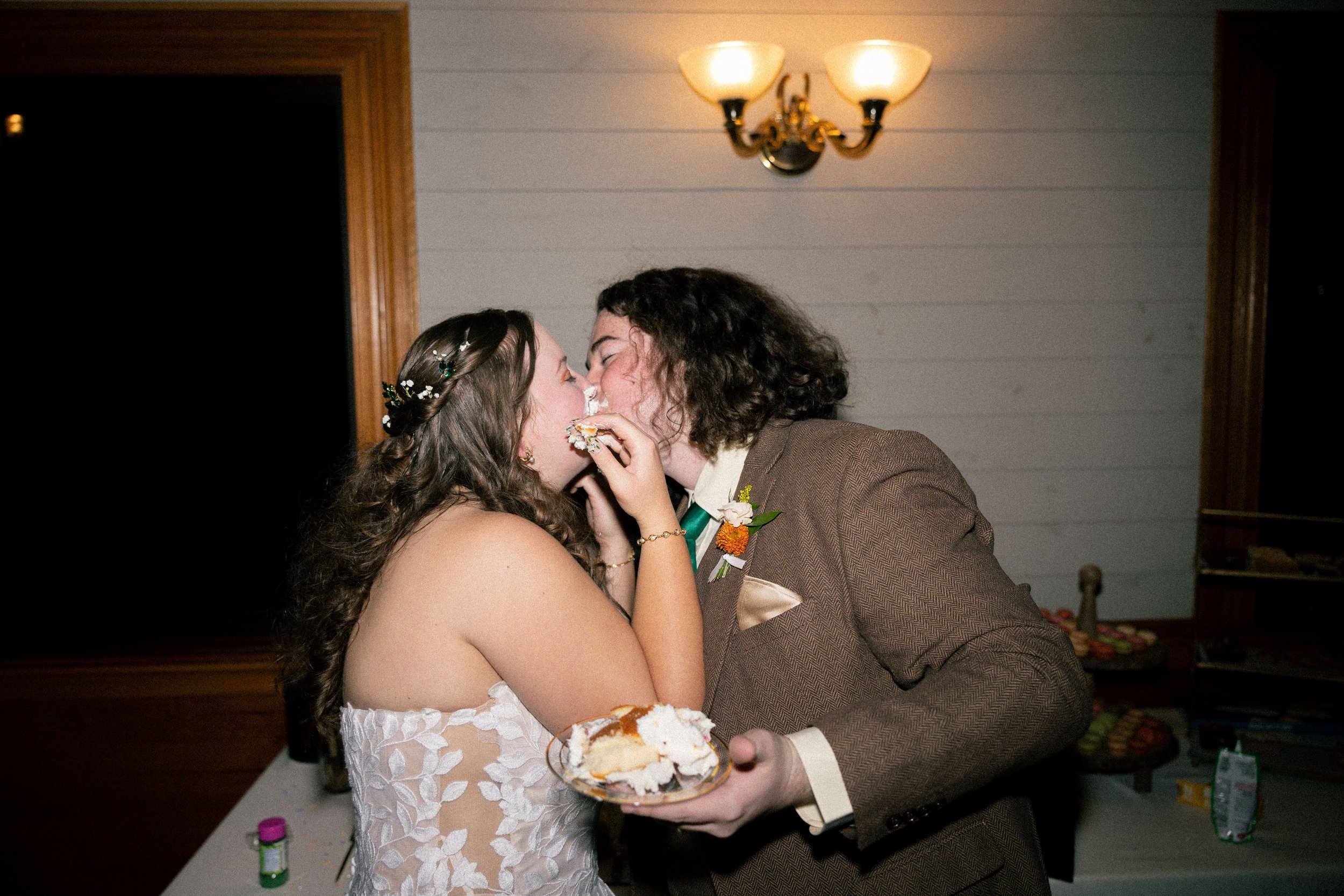 A bride and groom sharing a piece of wedding cake, kissing each other, in a warmly lit room.