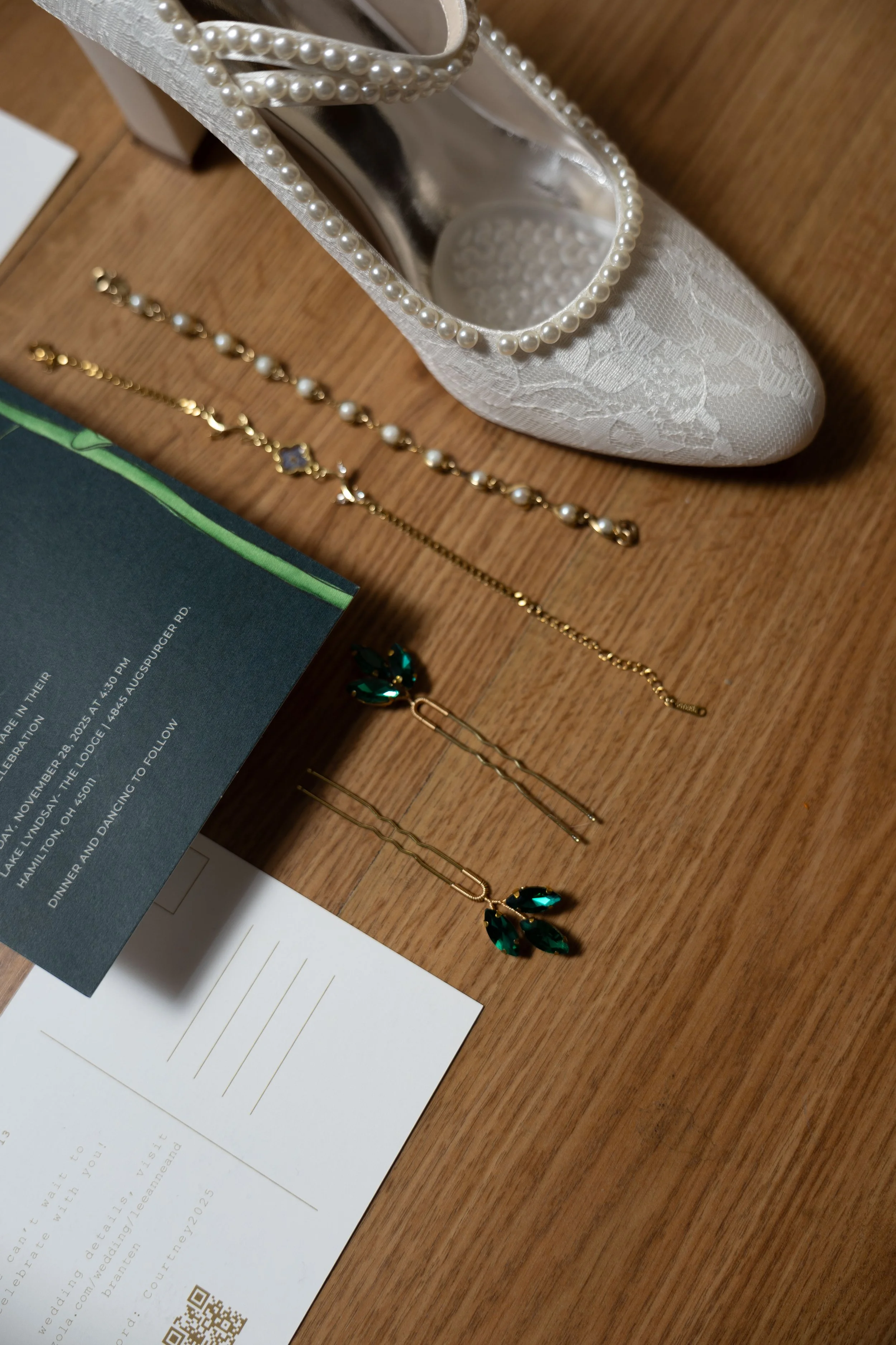White lace wedding shoe decorated with pearls, alongside gold and pearl bracelets, green gemstone earrings, and wedding invitation cards on wooden surface.