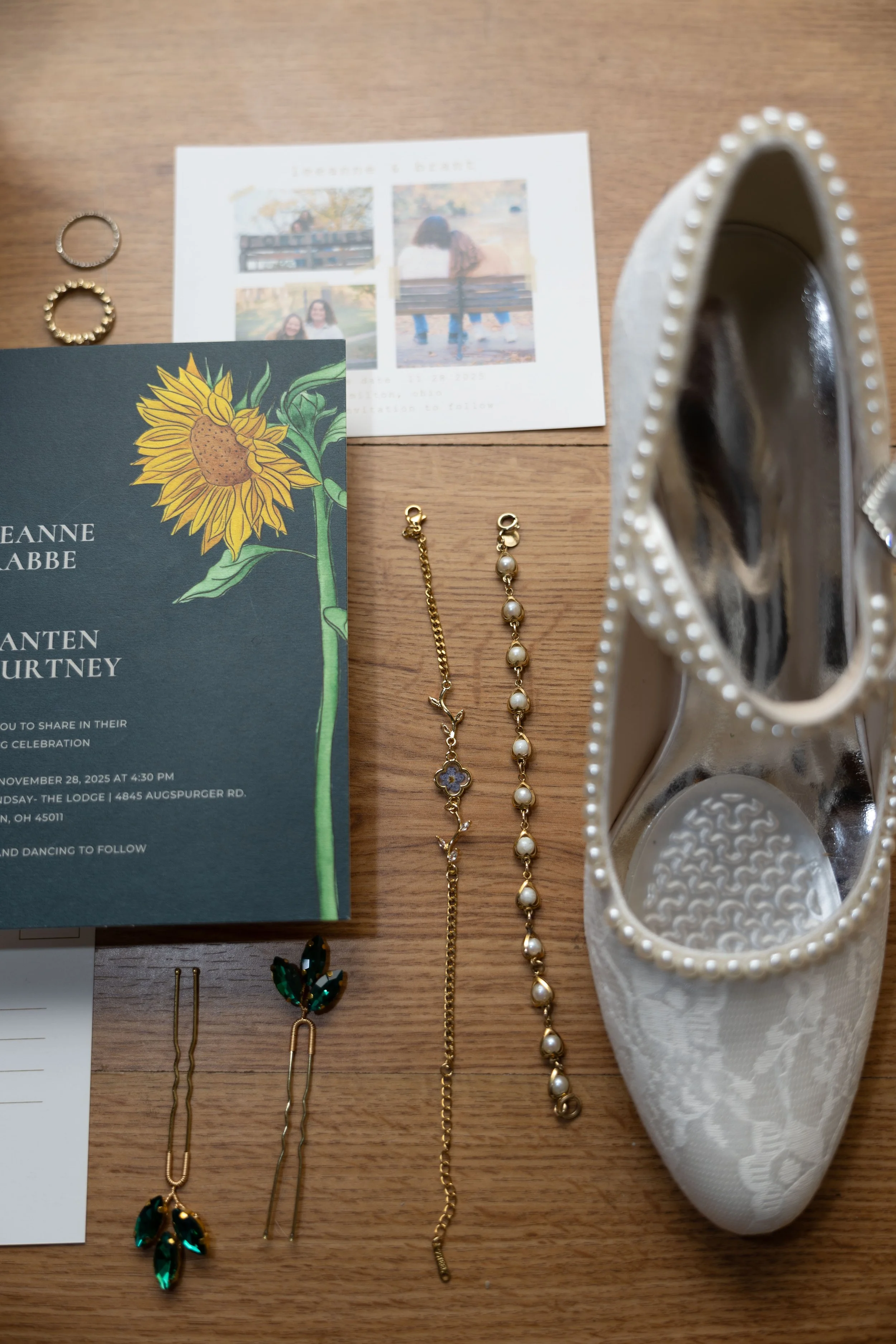 A collection of jewelry and event invitations on a wooden surface, including pearl and beaded bracelets, earrings with green gemstones and pearls, a white floral-patterned high-heeled shoe, a wedding invitation, and a photo card with four pictures of