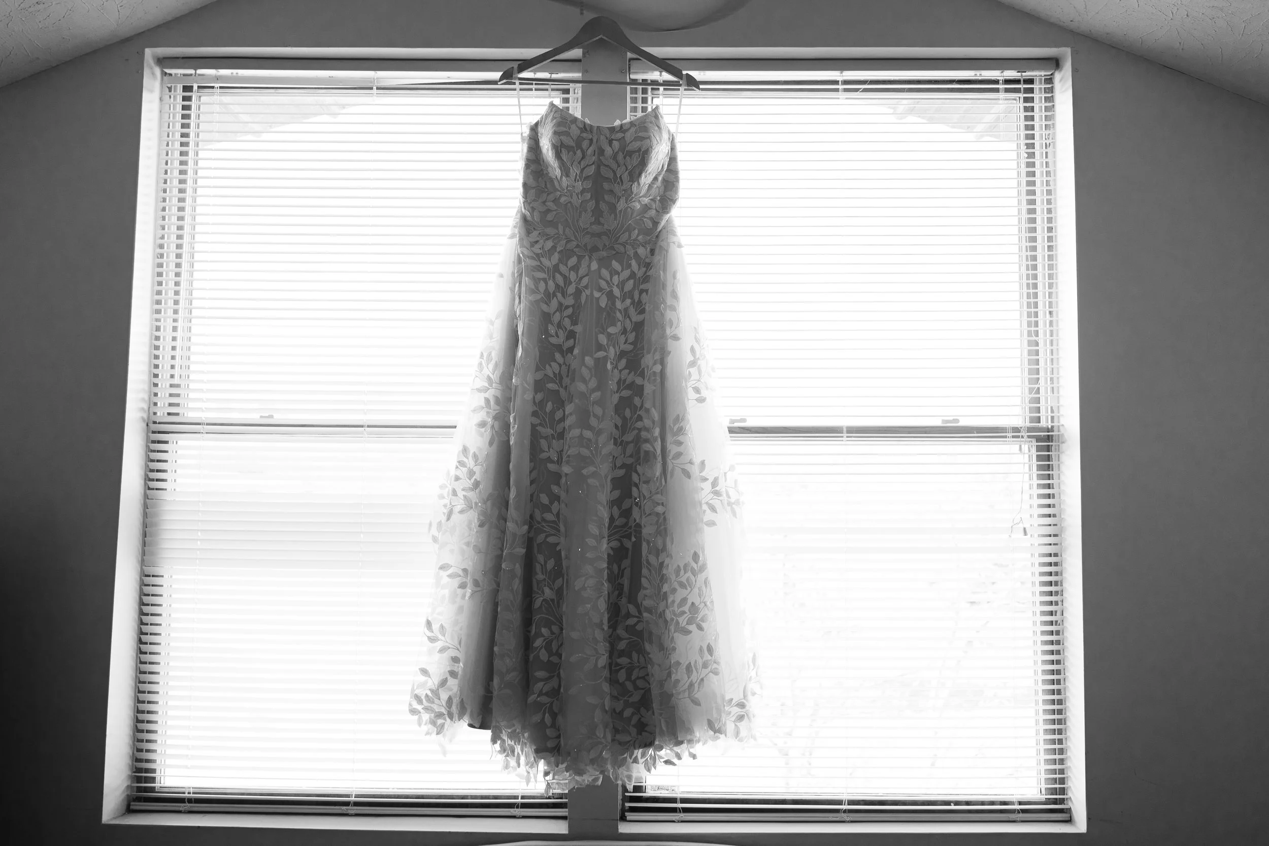 A wedding dress hanging on a hanger in front of a window with blinds.