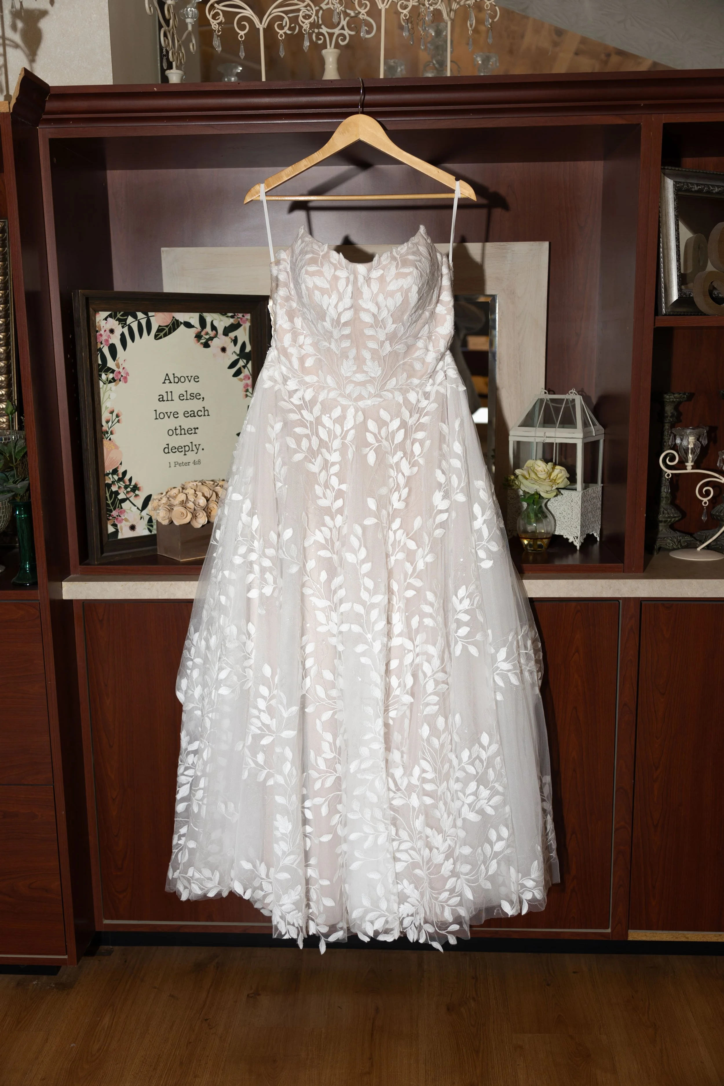 White wedding dress with floral embroidery hanging on a wooden hanger in front of a wooden cabinet.