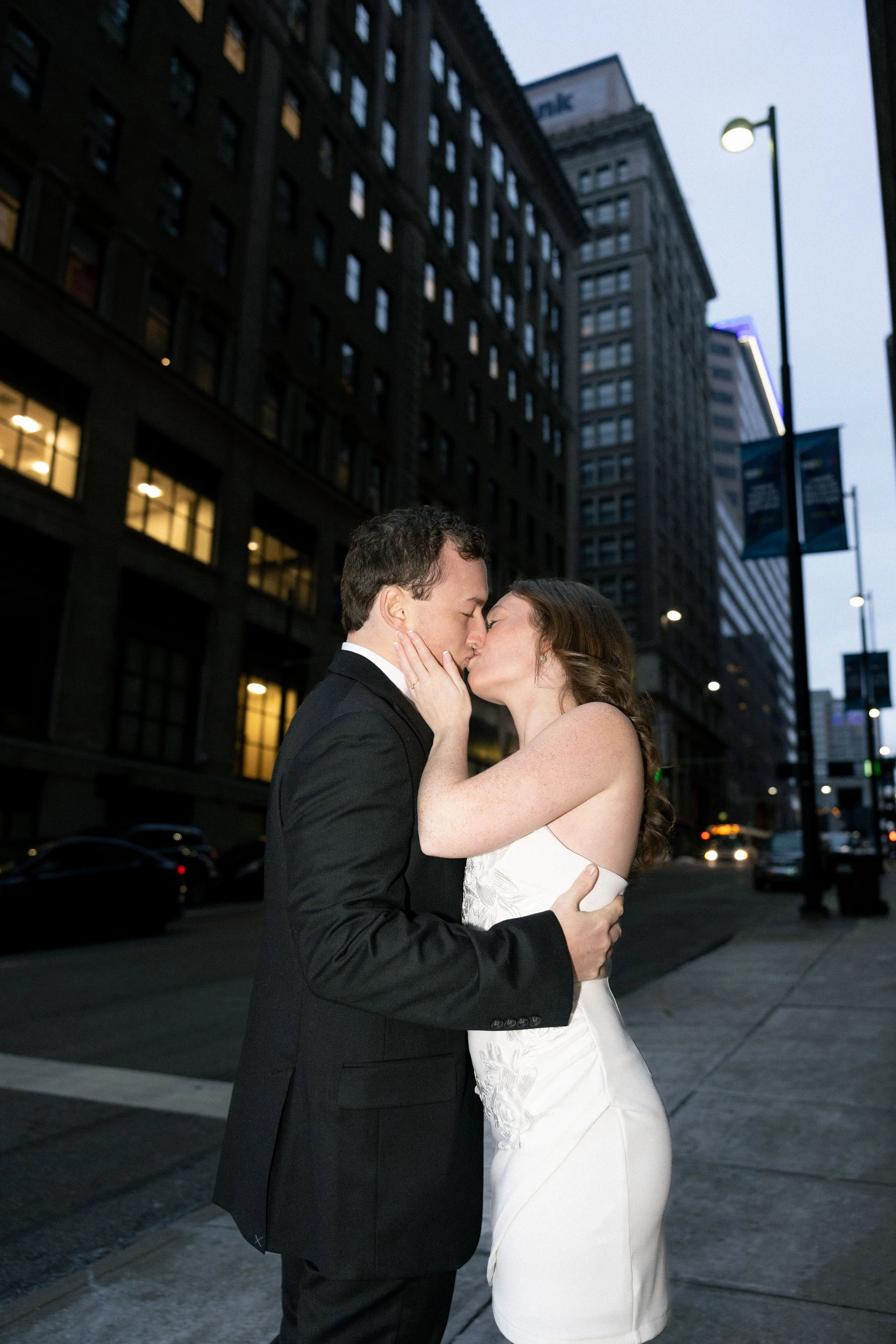 A couple in wedding attire sharing a kiss on a city street at dusk, with tall buildings and streetlights in the background.