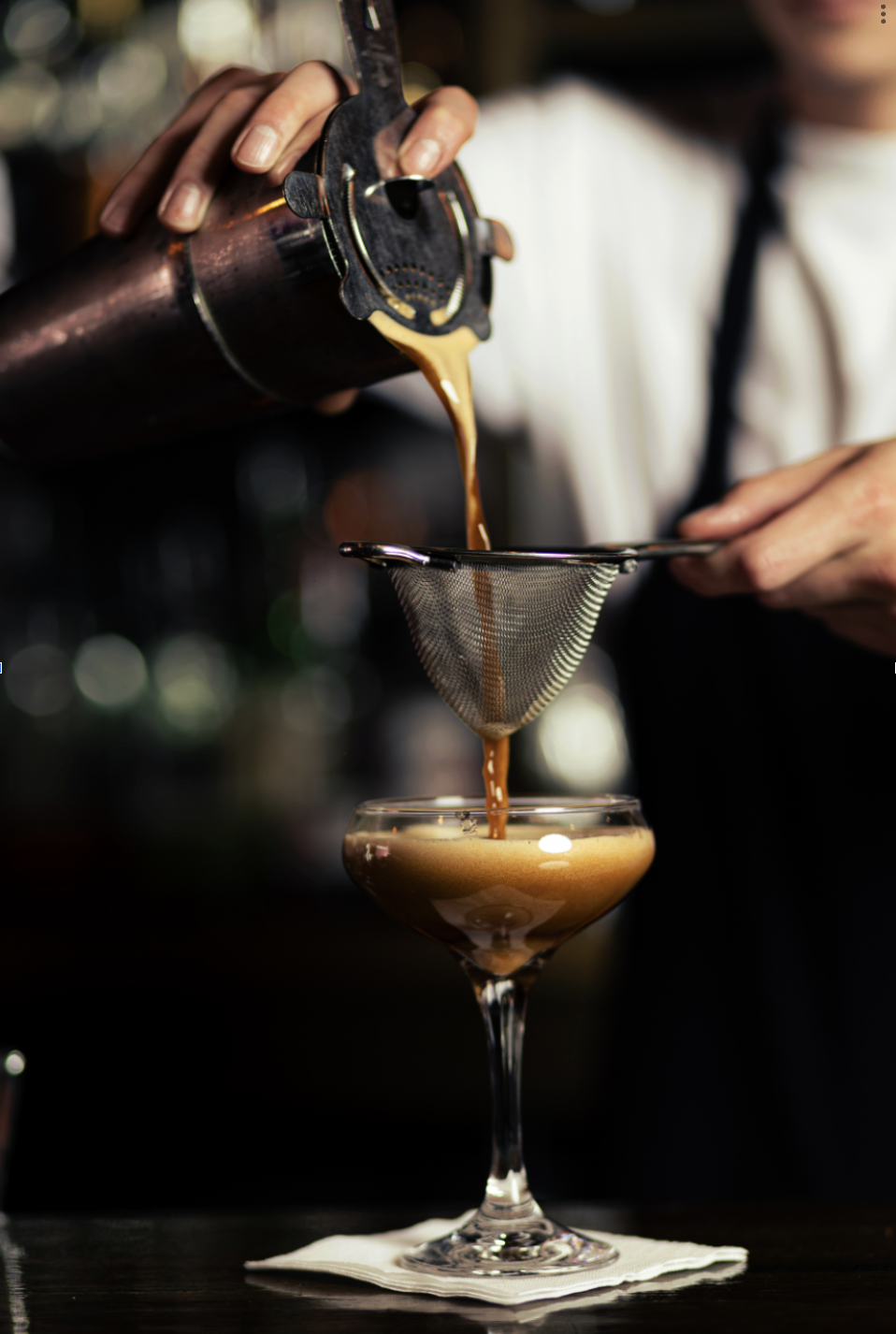 A bartender pours a freshly made espresso martini through a strainer into a coupe glass, with a dark, blurred bar background.