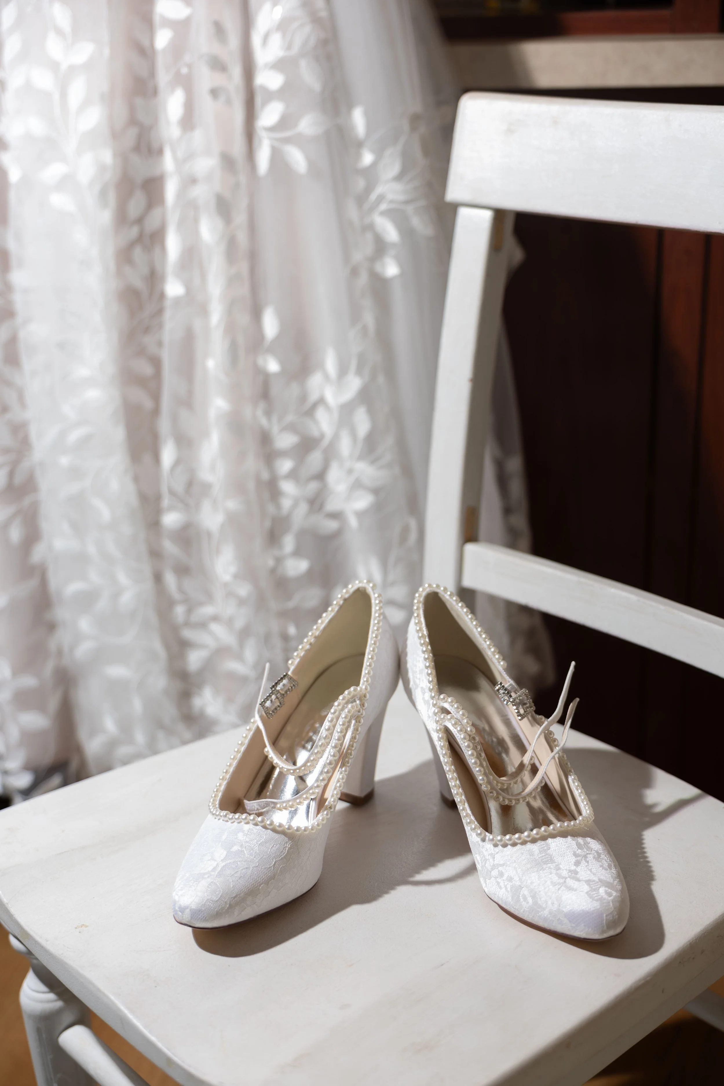 A pair of elegant white wedding shoes with lace pattern and pearl decorations placed on a white chair near a window with sheer leafy-patterned curtains.