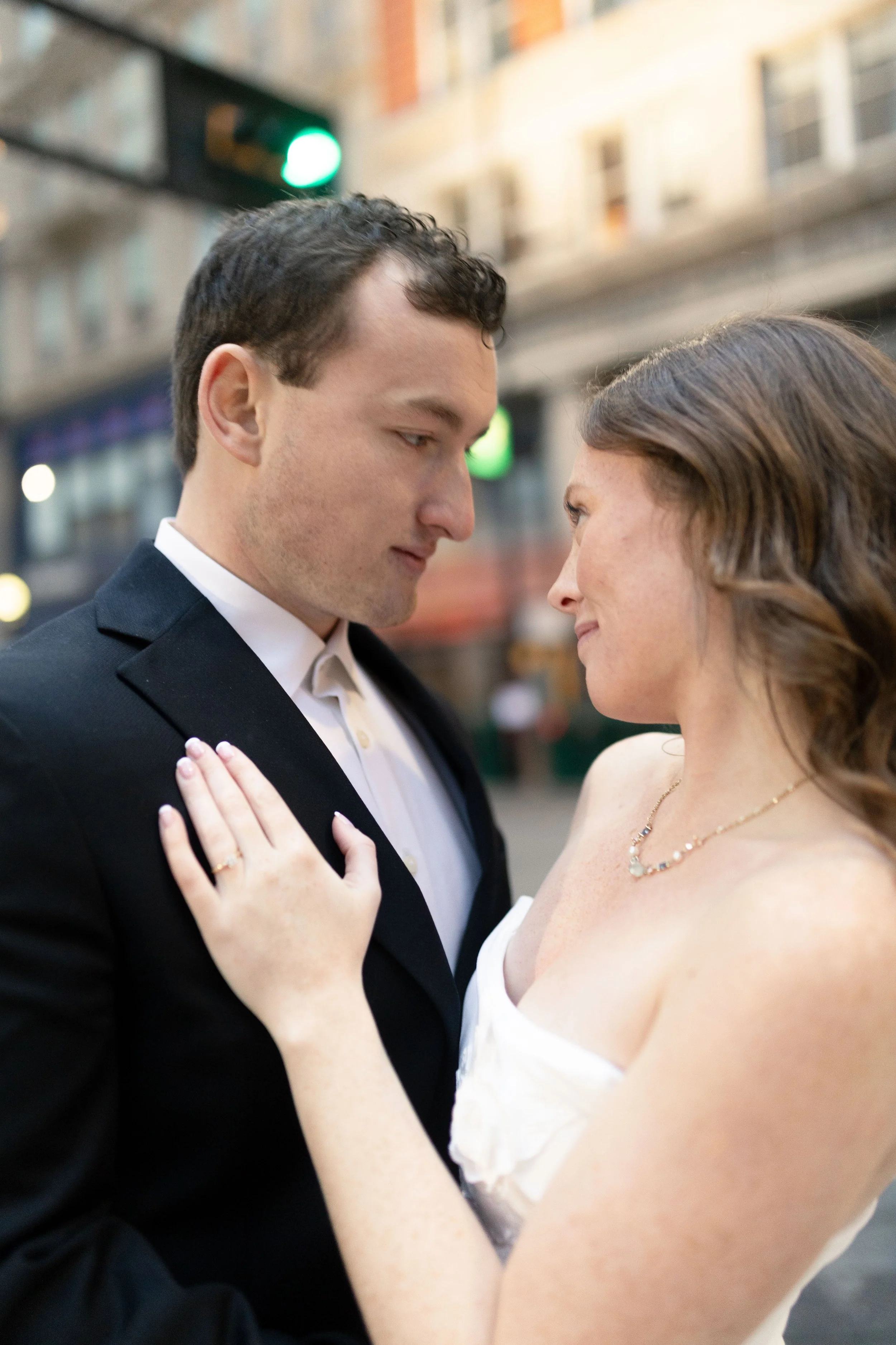 A couple dressed in formal attire sharing an intimate moment on a city street at dusk, with buildings and traffic lights in the background.