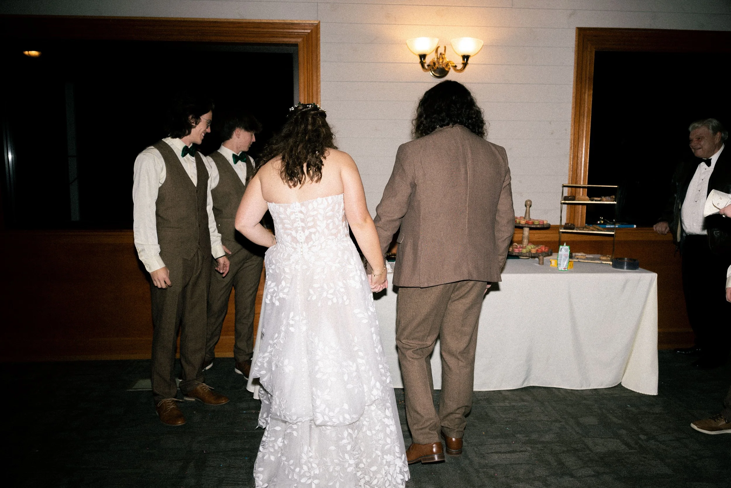 A bride and groom with their backs turned to the camera, standing at a table with desserts, while three groomsmen stand nearby at a wedding reception.