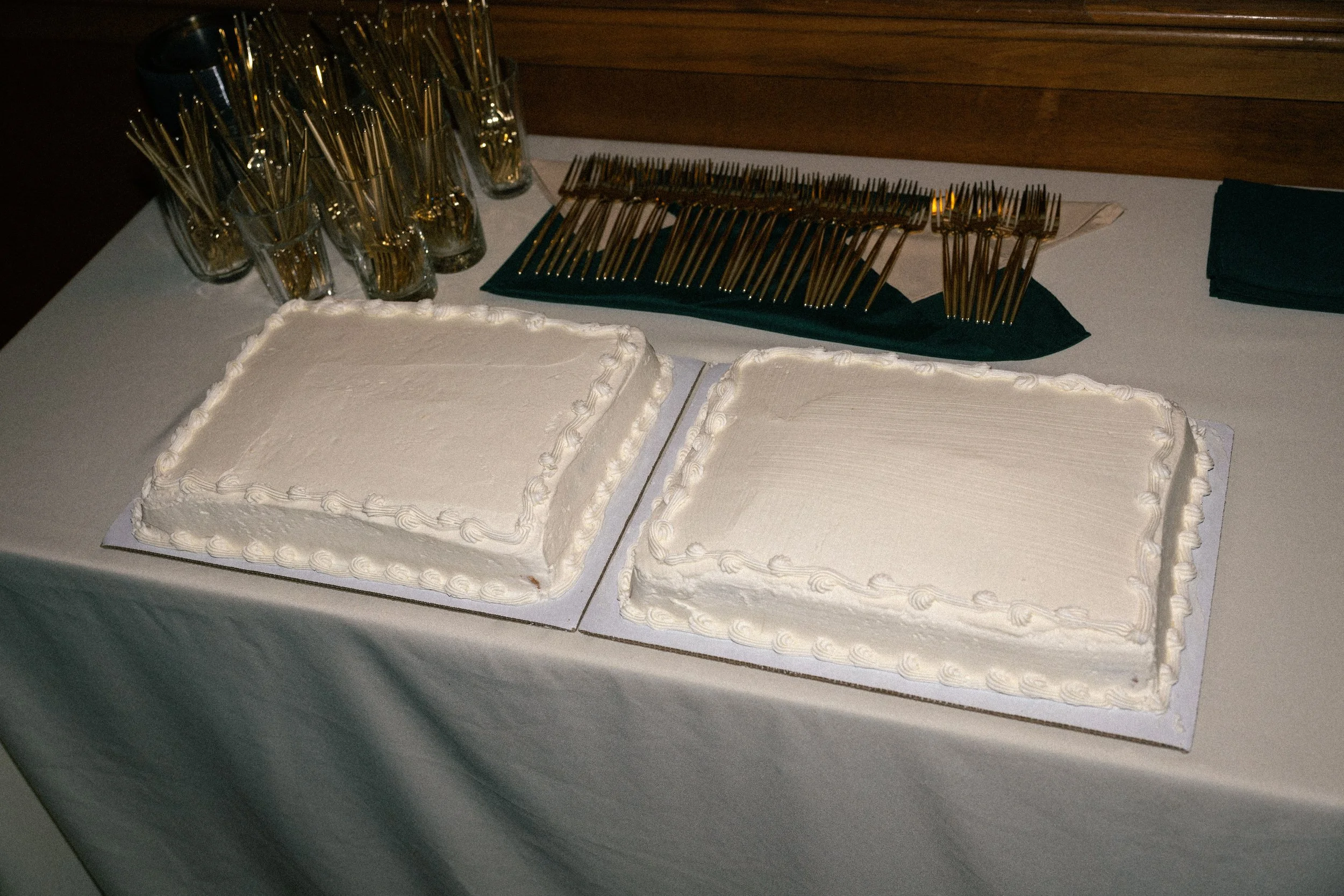 Two rectangular cakes with white frosting on a table, with gold forks and glasses arranged behind them.