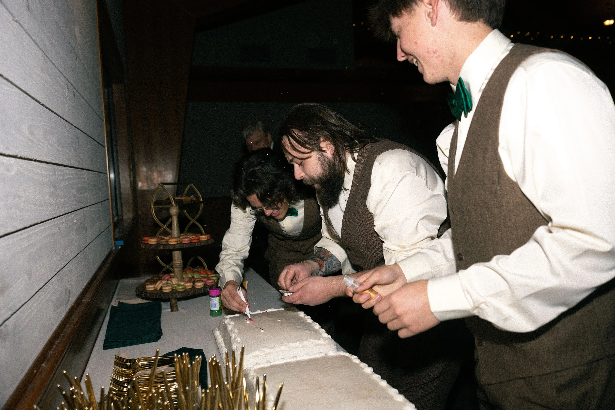 Group of three people in vintage clothing decorating a wedding cake with white frosting at a celebration, with macaron desserts and decorative items on the table.