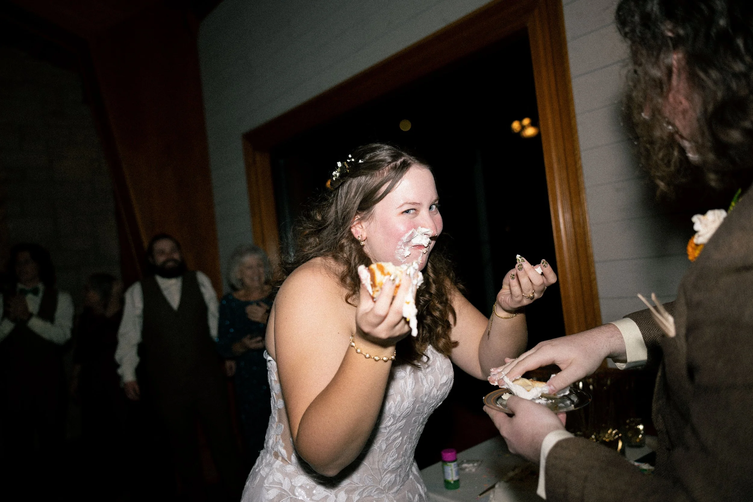 A young woman in a white dress with a floral pattern is at a wedding reception, smiling as she eats a piece of cake. Her face and nose are covered in white icing. She is holding cake in her left hand and a plate with more cake in her right hand, whil
