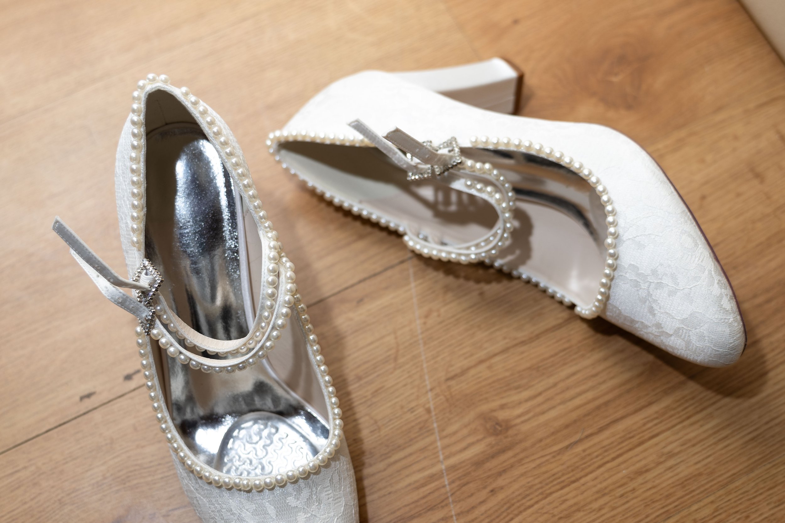 A pair of white bridal shoes with pearl embellishments on a wooden floor, with the left shoe showing an internal mirror.