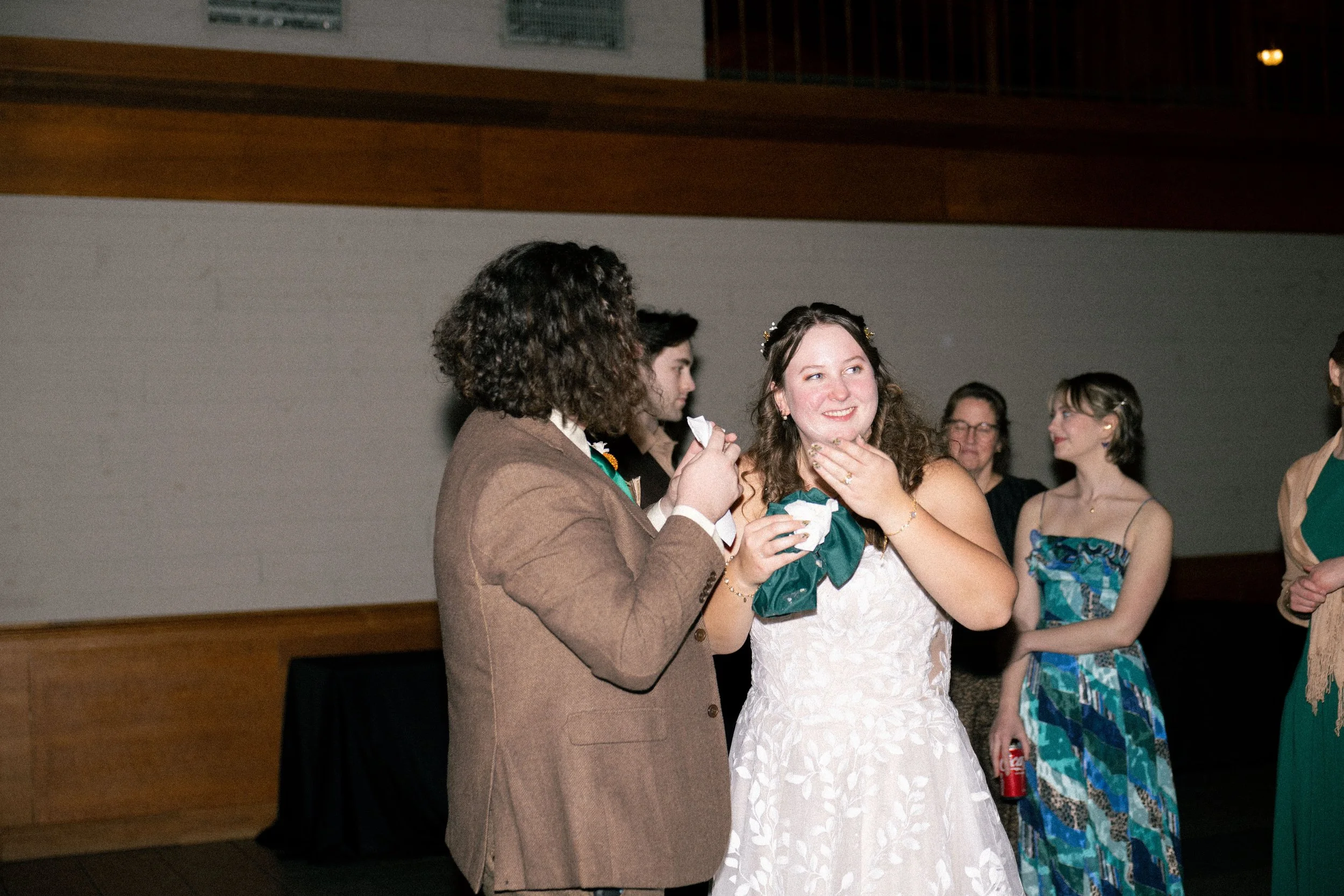 Group of people at a social event, with a woman in a white lace dress smiling and touching her chin, surrounded by others in formal attire.