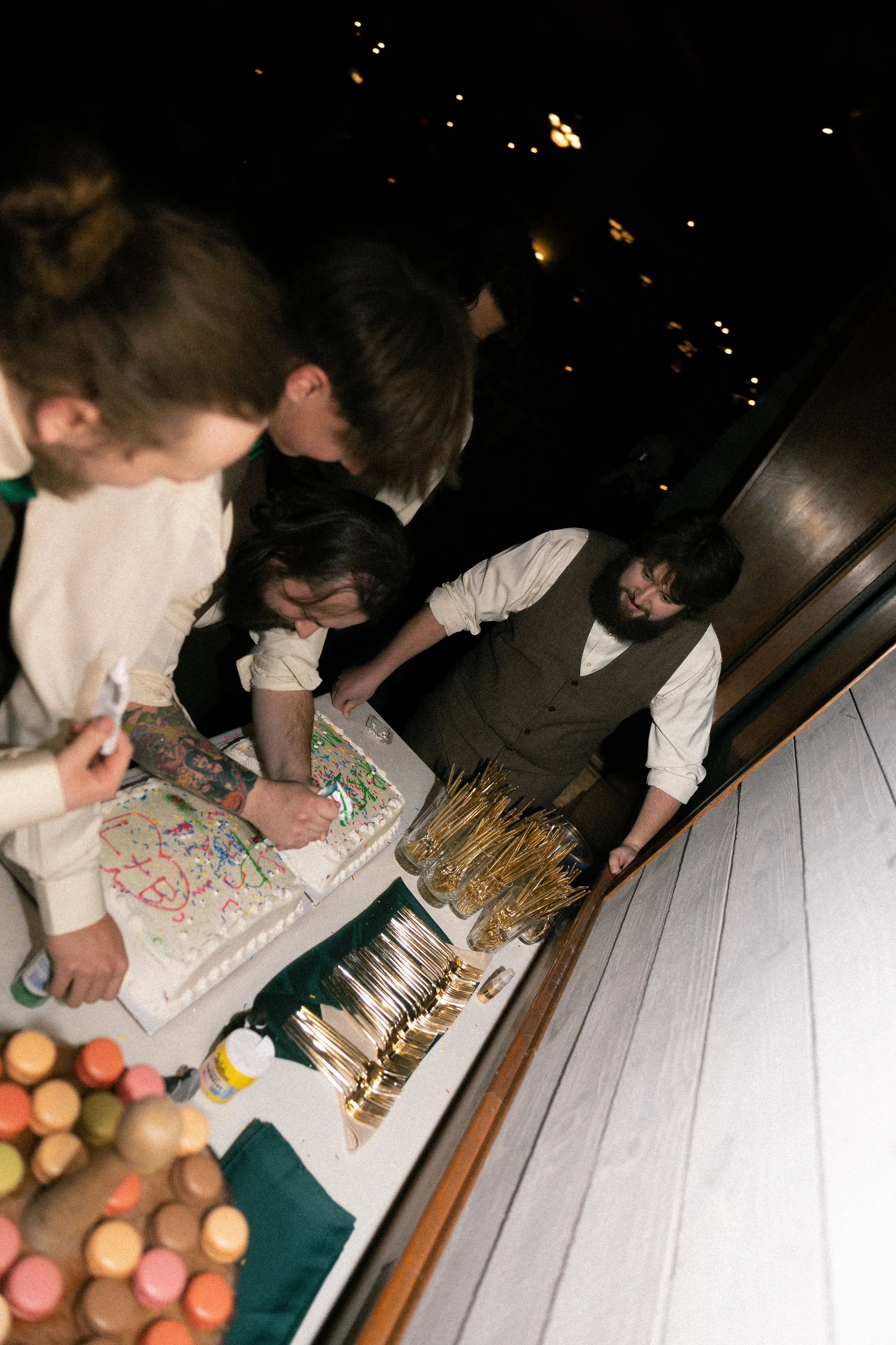 Group of people celebrating a birthday, cutting a decorated cake at a party with a dark background and warm lighting.