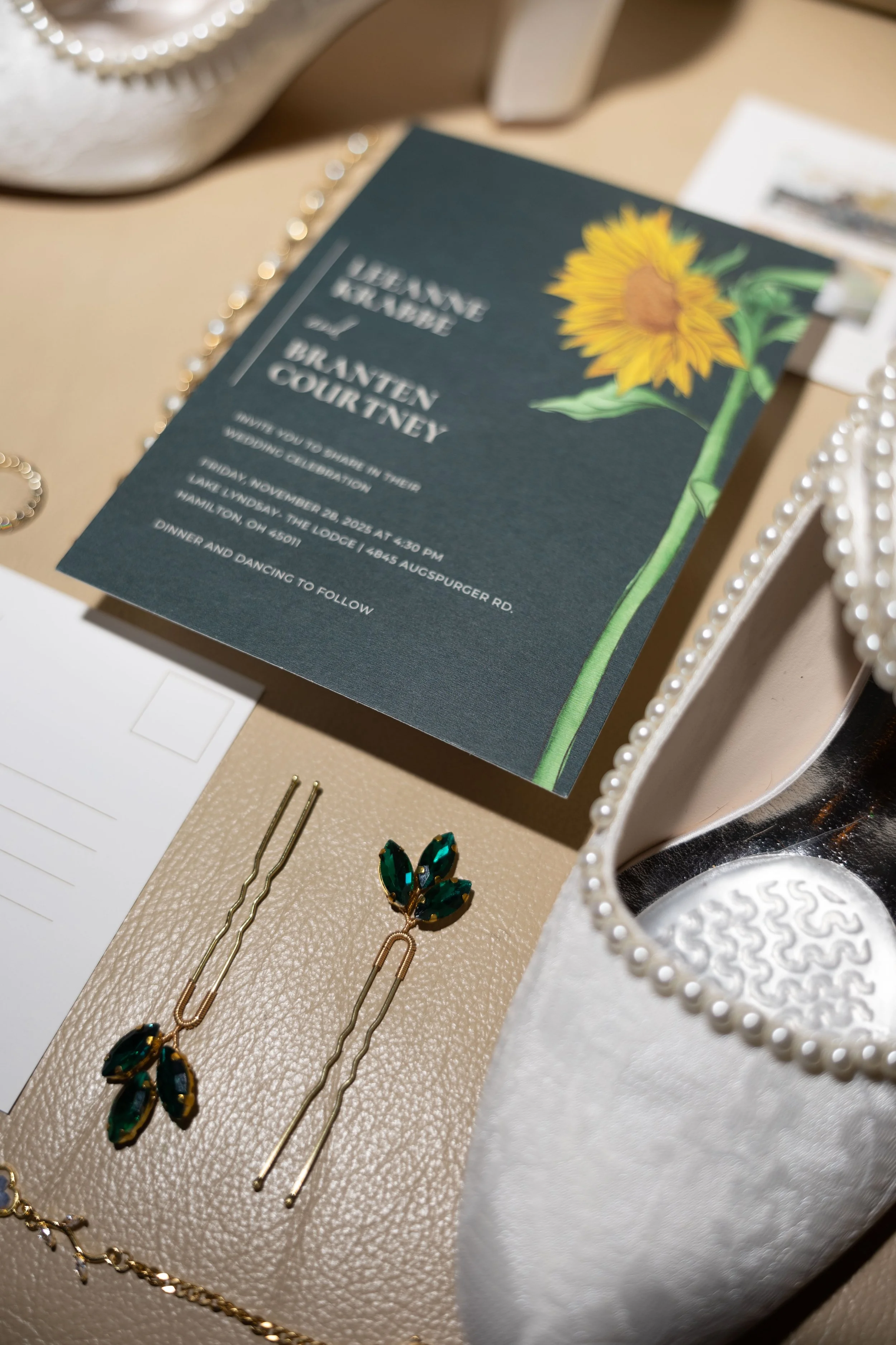 Wedding invitation with a sunflower design, a pair of green gemstone earrings, and a white pearl-decorated mirror.