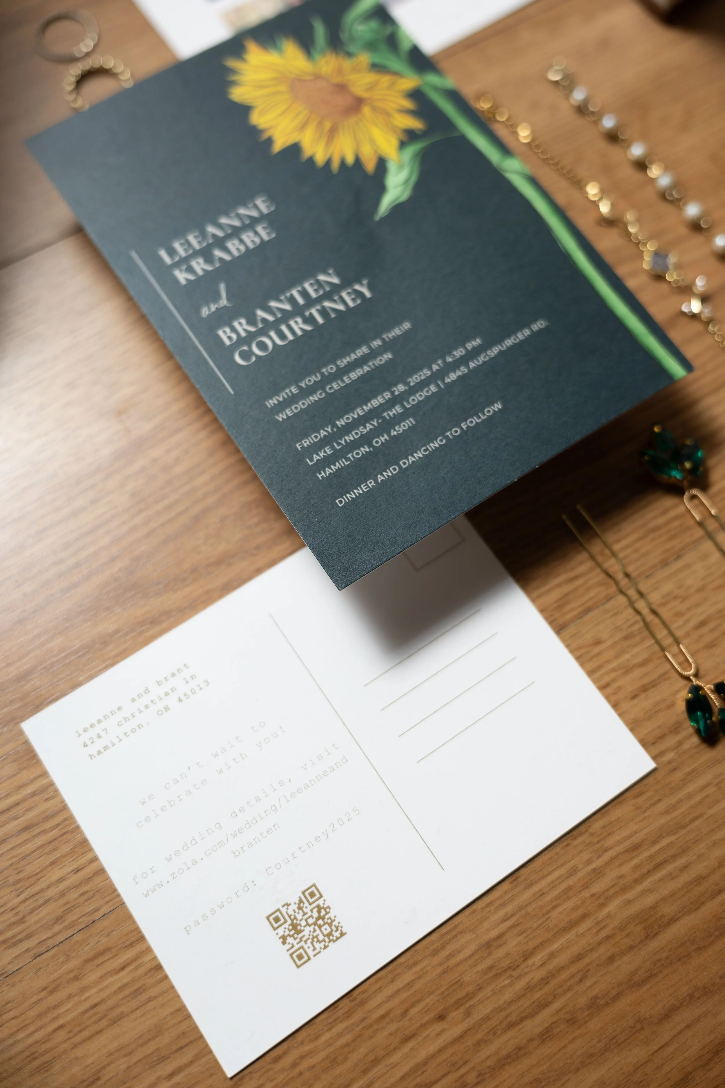 Wedding invitation card for Leanne Krabbe and Brantney Courtney with sunflower graphic, event details, and a postcard with RSVP information and a QR code, surrounded by jewelry items.
