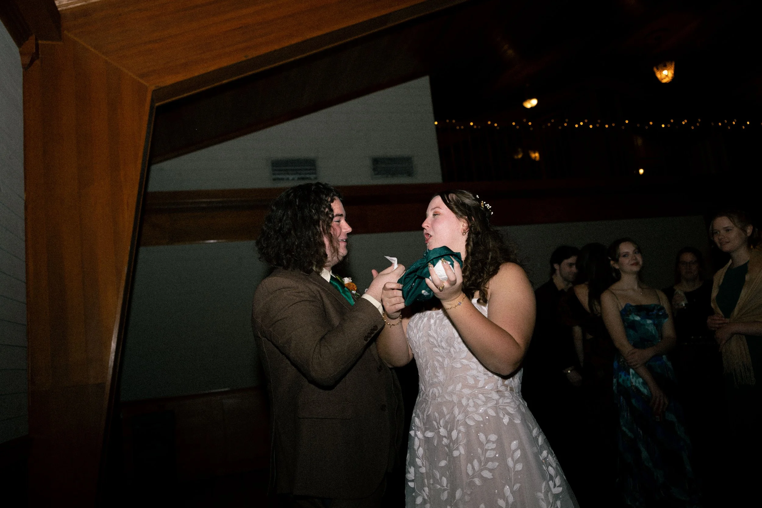 A couple sharing a moment at a wedding reception with guests in the background, one in a brown suit and the other in a white wedding dress with leaf patterns.