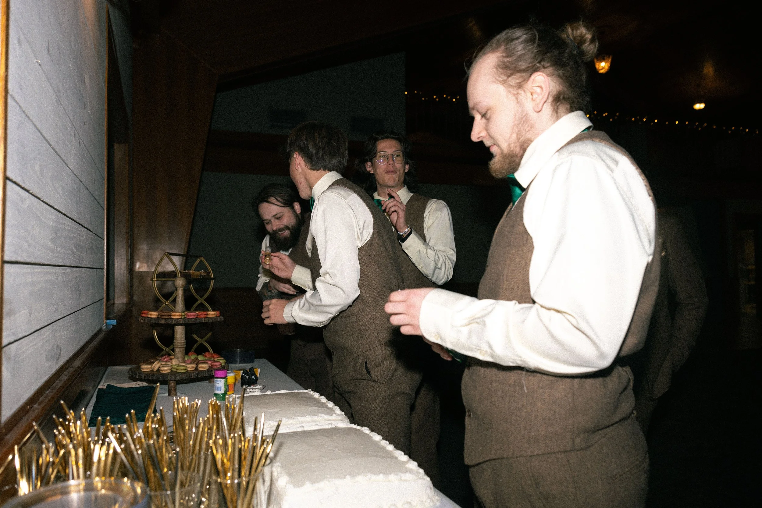 Four men in vintage-style clothing gathered around a table with desserts and decorations, possibly at a party or celebration, in a dimly lit room.