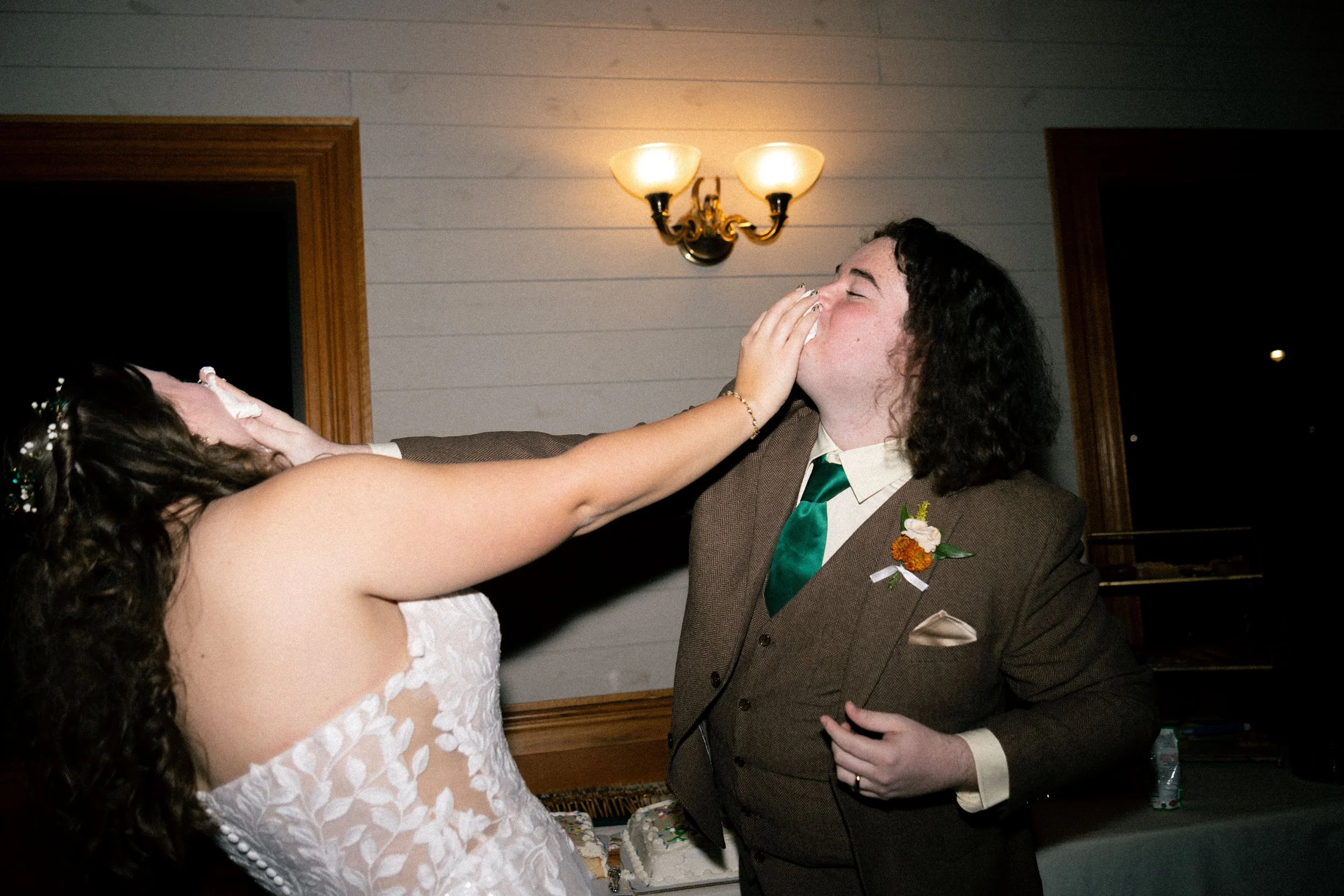 A woman in a wedding dress is playfully pretending to hit a man with a tissue while he smiles and covers his face in surprise, in a warmly lit room with a wall sconce.