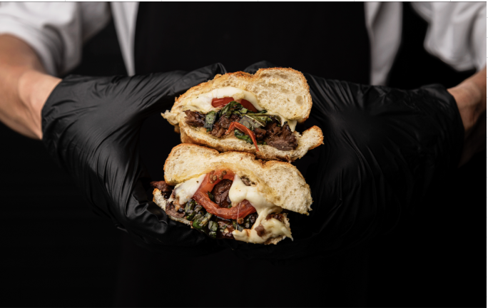 Person wearing black gloves holding a sliced sandwich with beef, tomatoes, greens, and cheese.