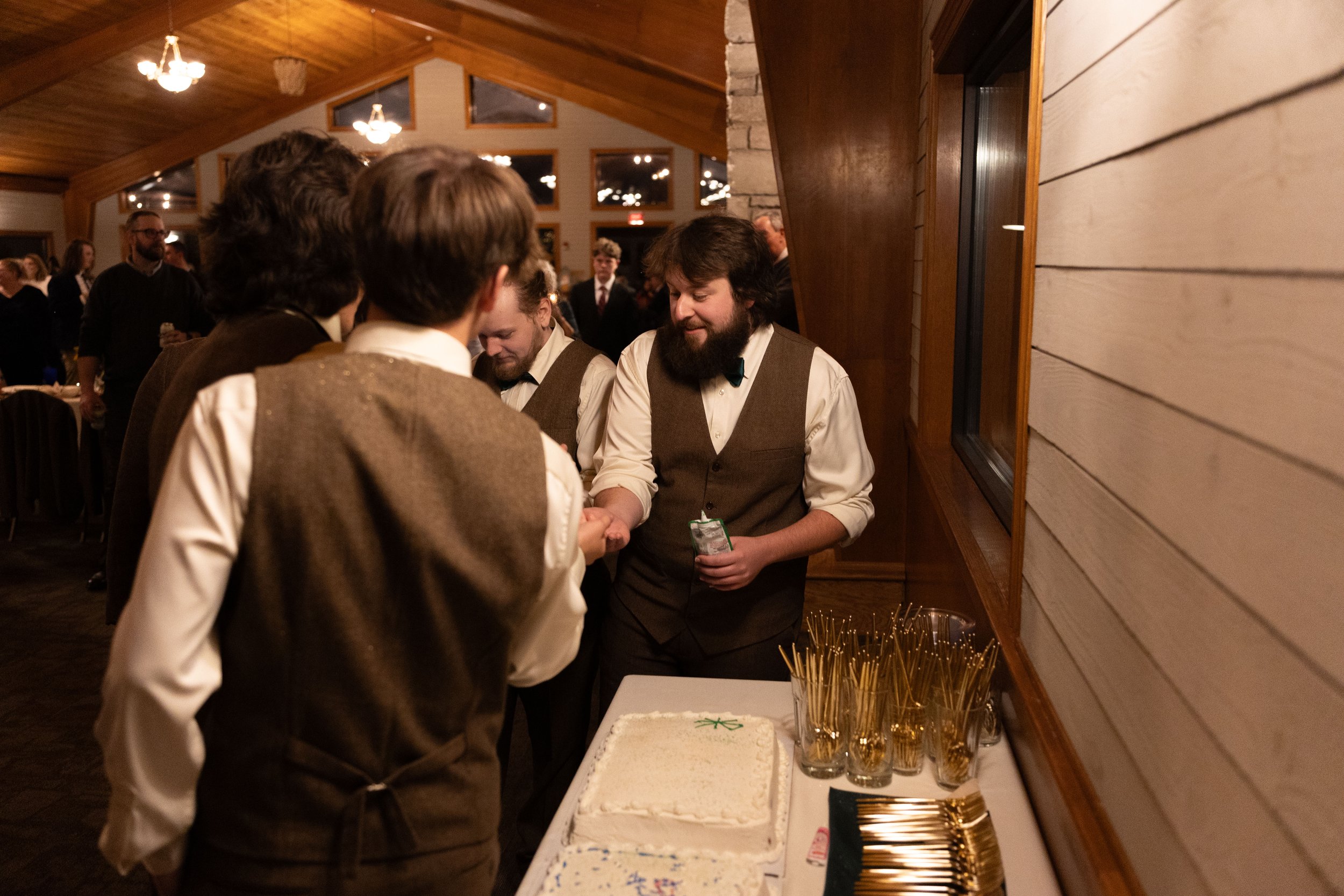 People in vintage-style attire gathered around a table with a cake at a cozy, wood-paneled indoor event, engaging in conversation and handshakes.