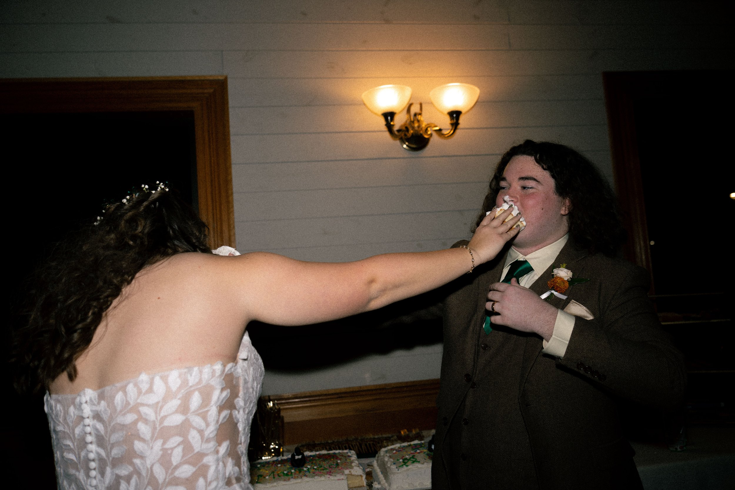 A woman in a white dress feeds a piece of cake to a man in a brown suit during a celebration in a room with wooden walls and a ceiling lamp.