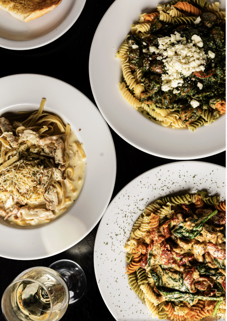Three plates of pasta dishes with different sauces and garnishes, a glass of white wine, and a piece of bread on a dark table.