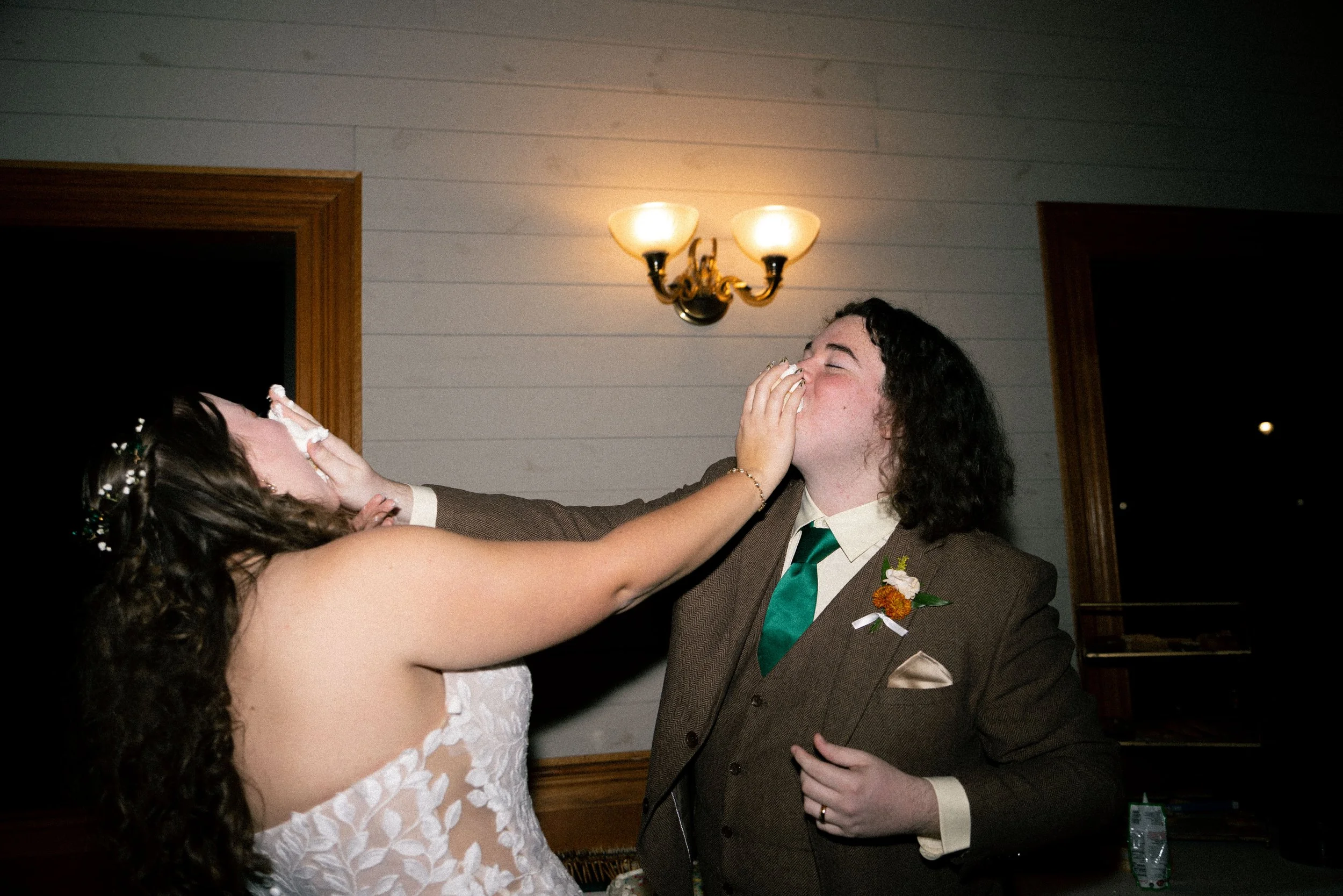 A woman playfully smears cake on a man's face during a wedding celebration, both are dressed in wedding attire, with the woman in a white lace dress and the man in a brown suit with a green tie, inside a room with wood trim and a wall-mounted light f