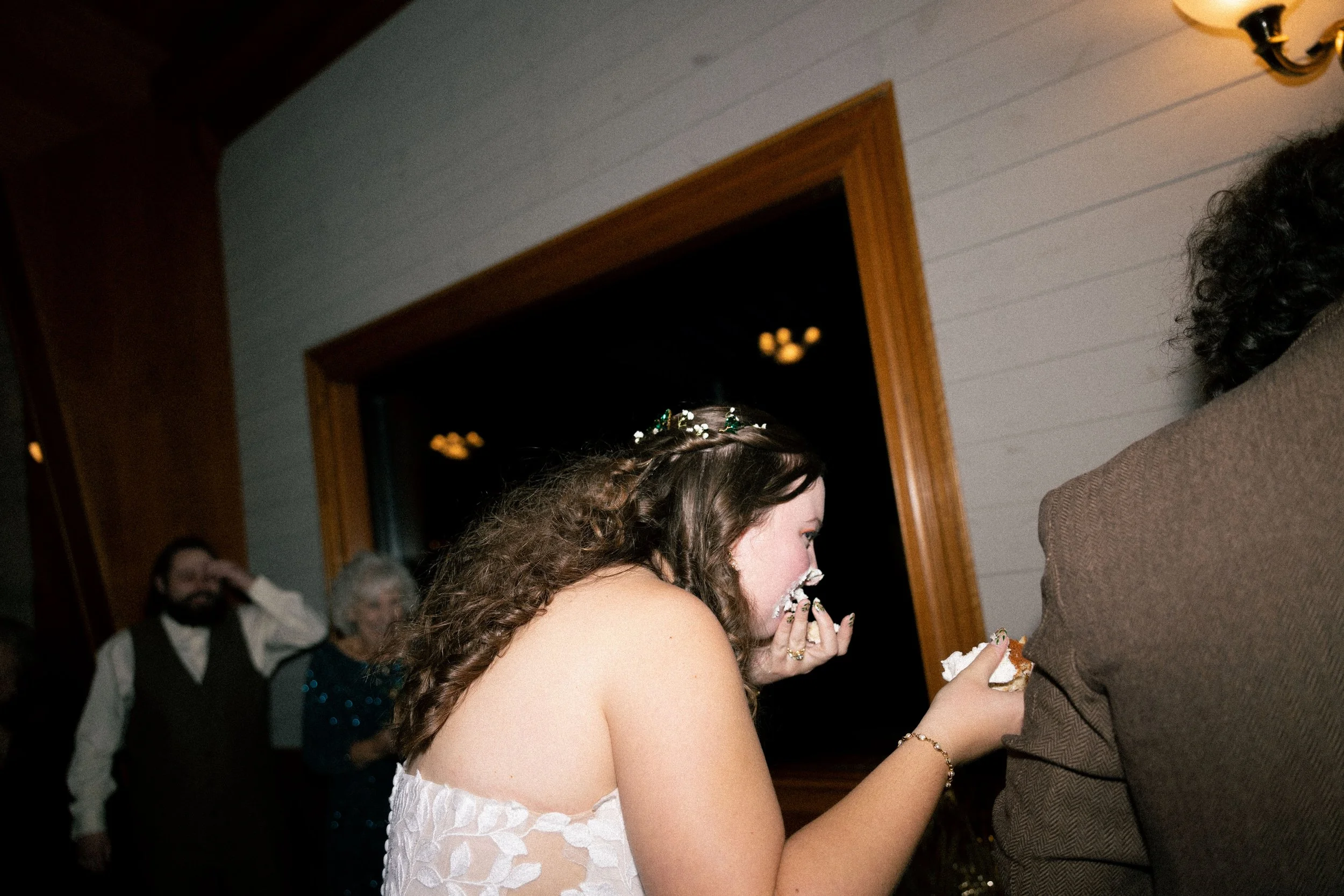 A woman in a white dress and a tiara is laughing while eating cake during her wedding reception, with guests visible in the background.