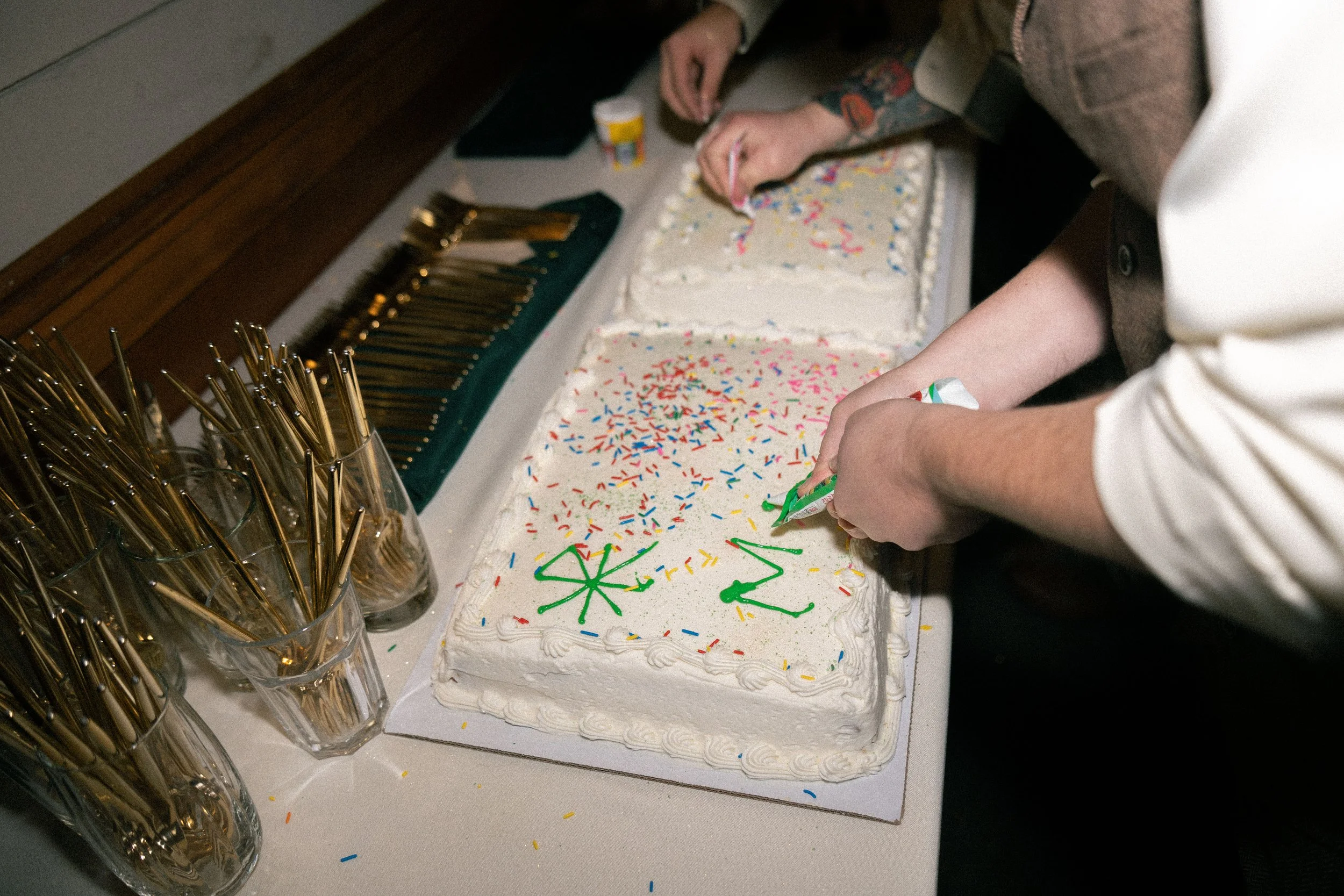 Person decorating a large rectangular birthday cake with green icing, colorful sprinkles, and the number '22' on top, using a piping bag, with other people present.