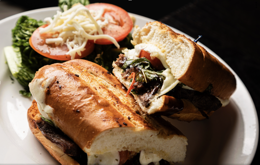 A close-up of a sandwich with cheese, vegetables, and meat, served with a side salad of sliced tomatoes, shredded cheese, and greens on a white plate.