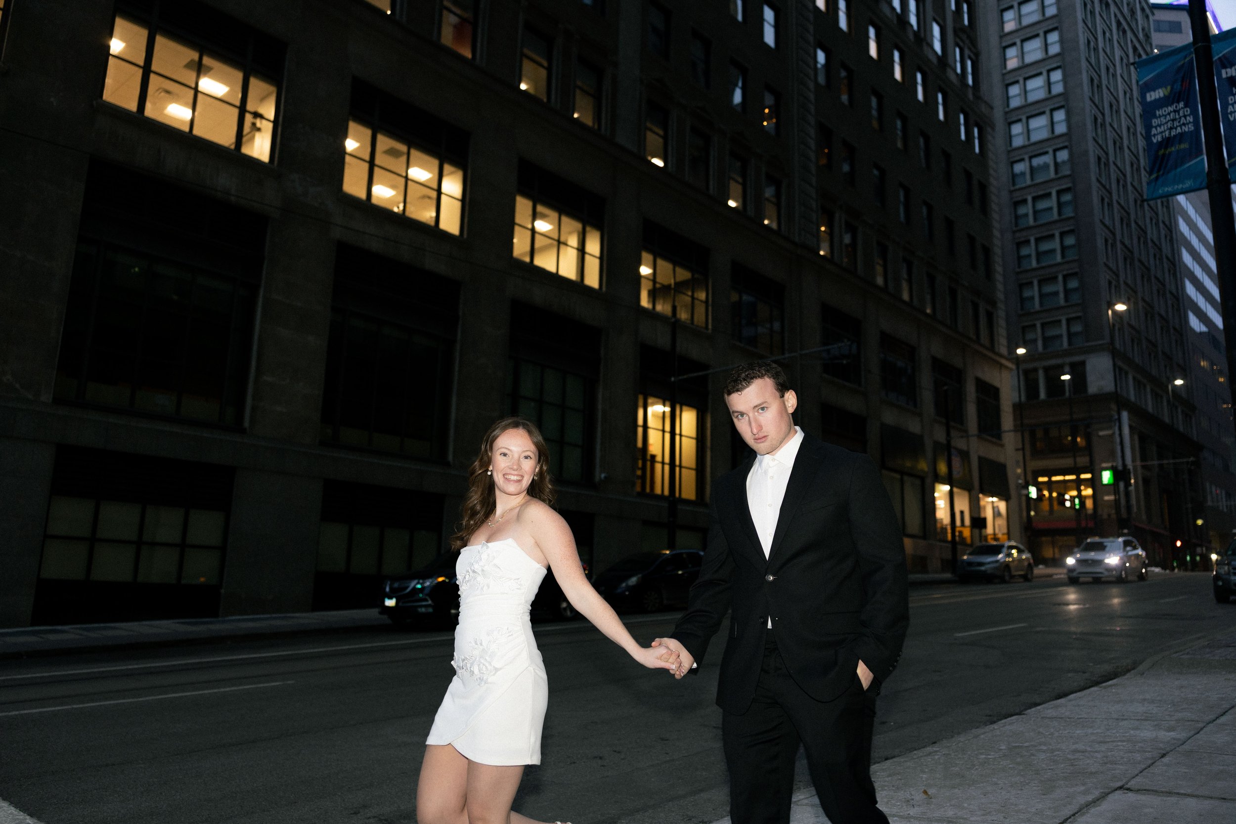 A young woman in a strapless white dress and a young man in a black suit walk hand in hand on city street at night, with tall buildings and cars in the background.