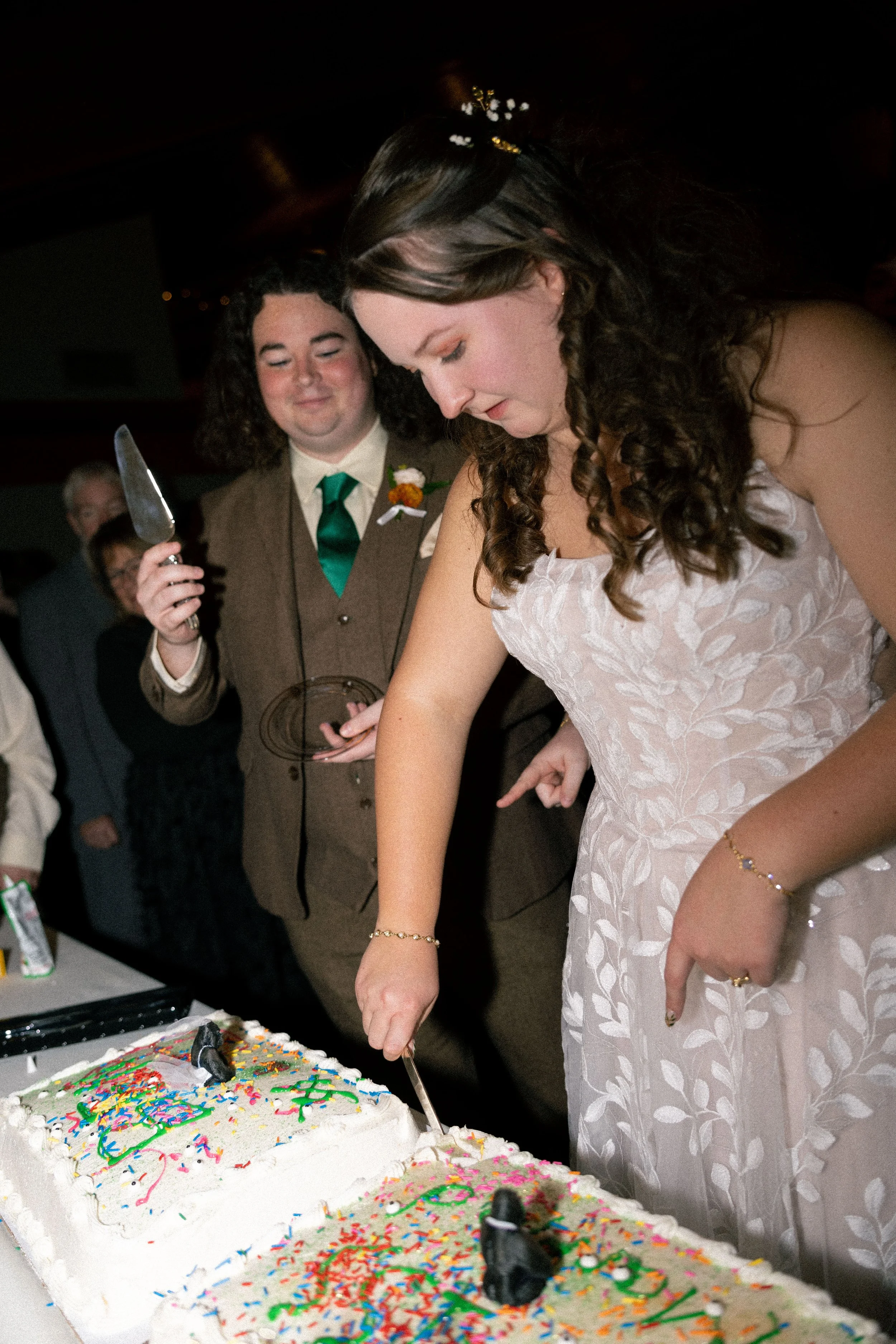 A woman in a white dress is cutting a cake at a celebration, with a man in a brown suit with a green tie standing nearby, holding a cake knife. They are surrounded by people in a festive atmosphere.