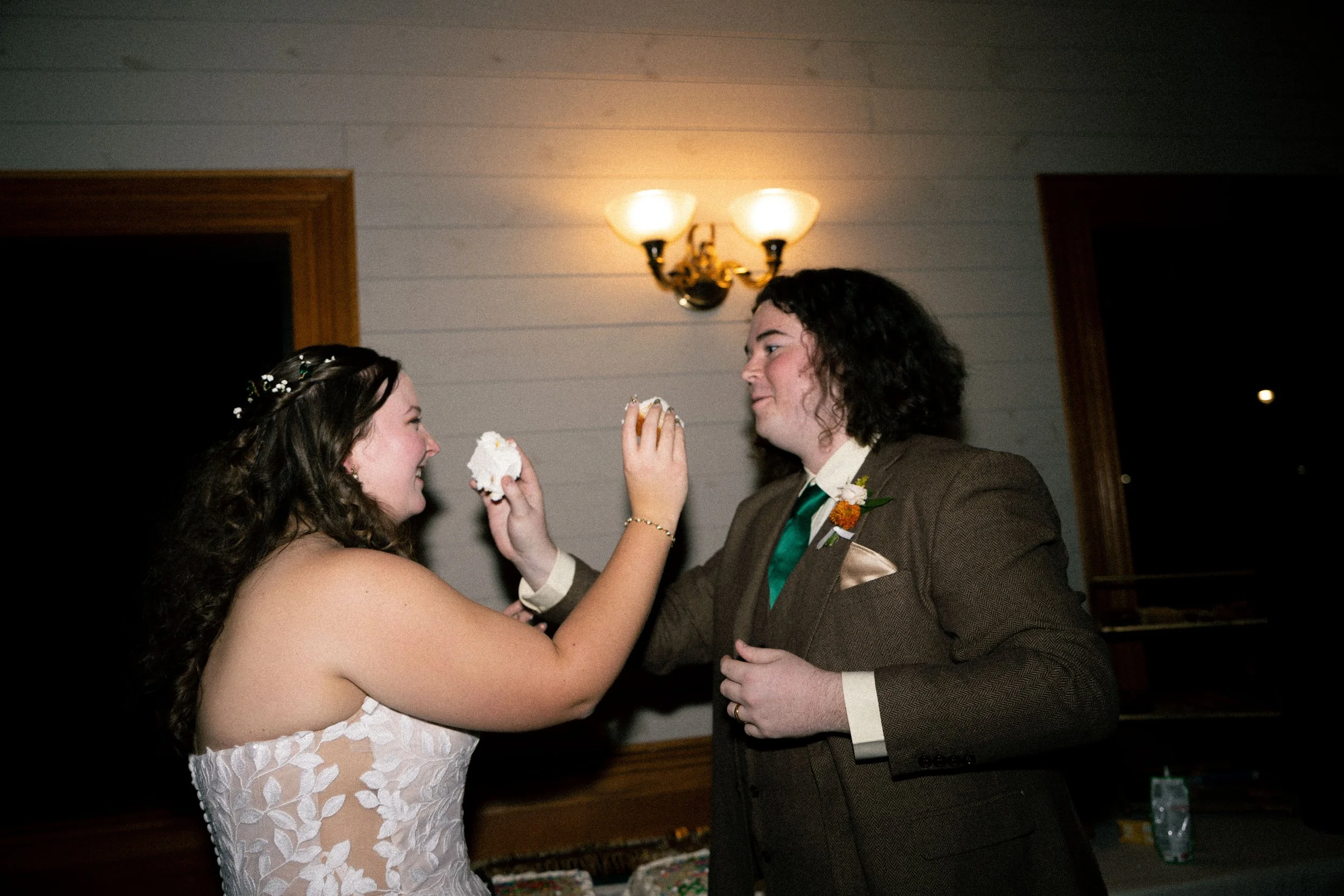 A bride and groom are sharing a joking cake face smear during their wedding reception. The bride is smiling, wearing a white lace dress with floral details and a floral headpiece. The groom is dressed in a brown suit with a green tie and boutonniere.