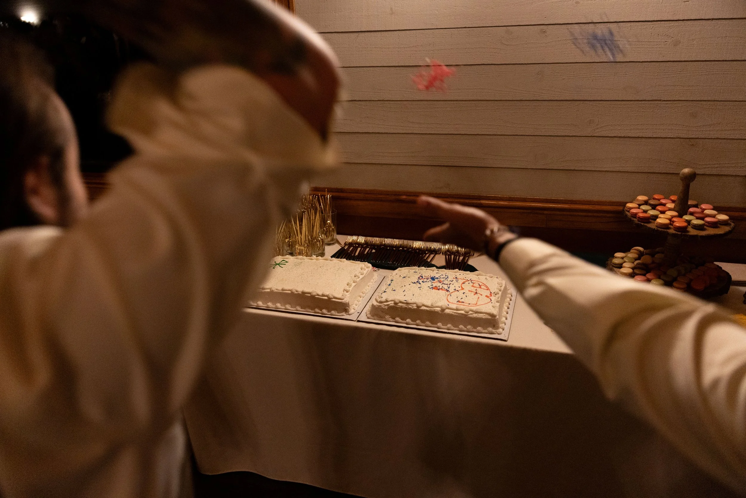 Two people celebrating with two cakes on a table, one with patriotic decorations, and a tiered tray with macarons in the background.
