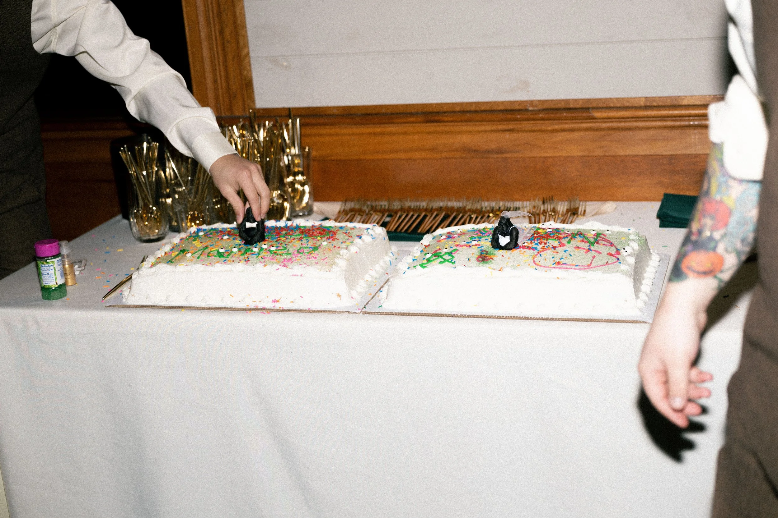 Two large rectangular birthday cakes decorated with multicolored sprinkles, with one person cutting the cake on the left and another person standing nearby on the right. There are gold utensils in glasses behind the cakes and small bottles of sprinkl